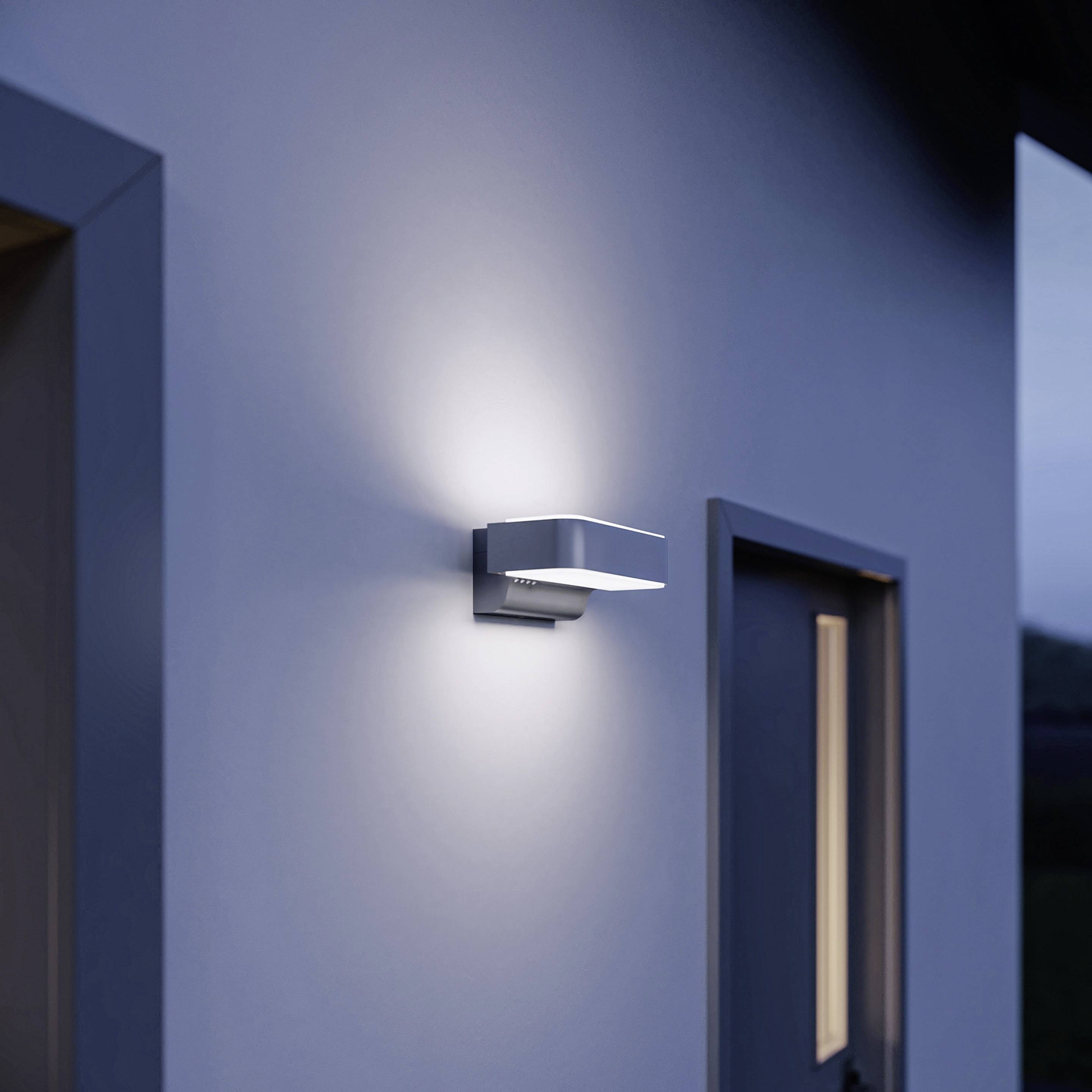 Moderne buitenwandlamp aan een witte muur naast een deur, verlicht boven en beneden. Verlicht de ingangszone 's nachts.