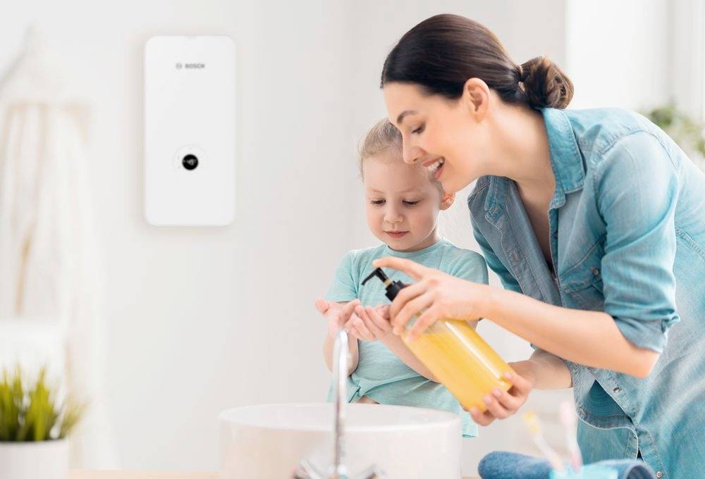 Een vrouw helpt een kind de handen te wassen met zeep bij een wasbak. Op de achtergrond is een waterreservoir zichtbaar.
