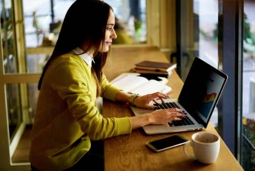 Een vrouw zit aan een tafel in een café en werkt geconcentreerd op een laptop. Naast haar liggen notitieboeken en een kopje koffie.