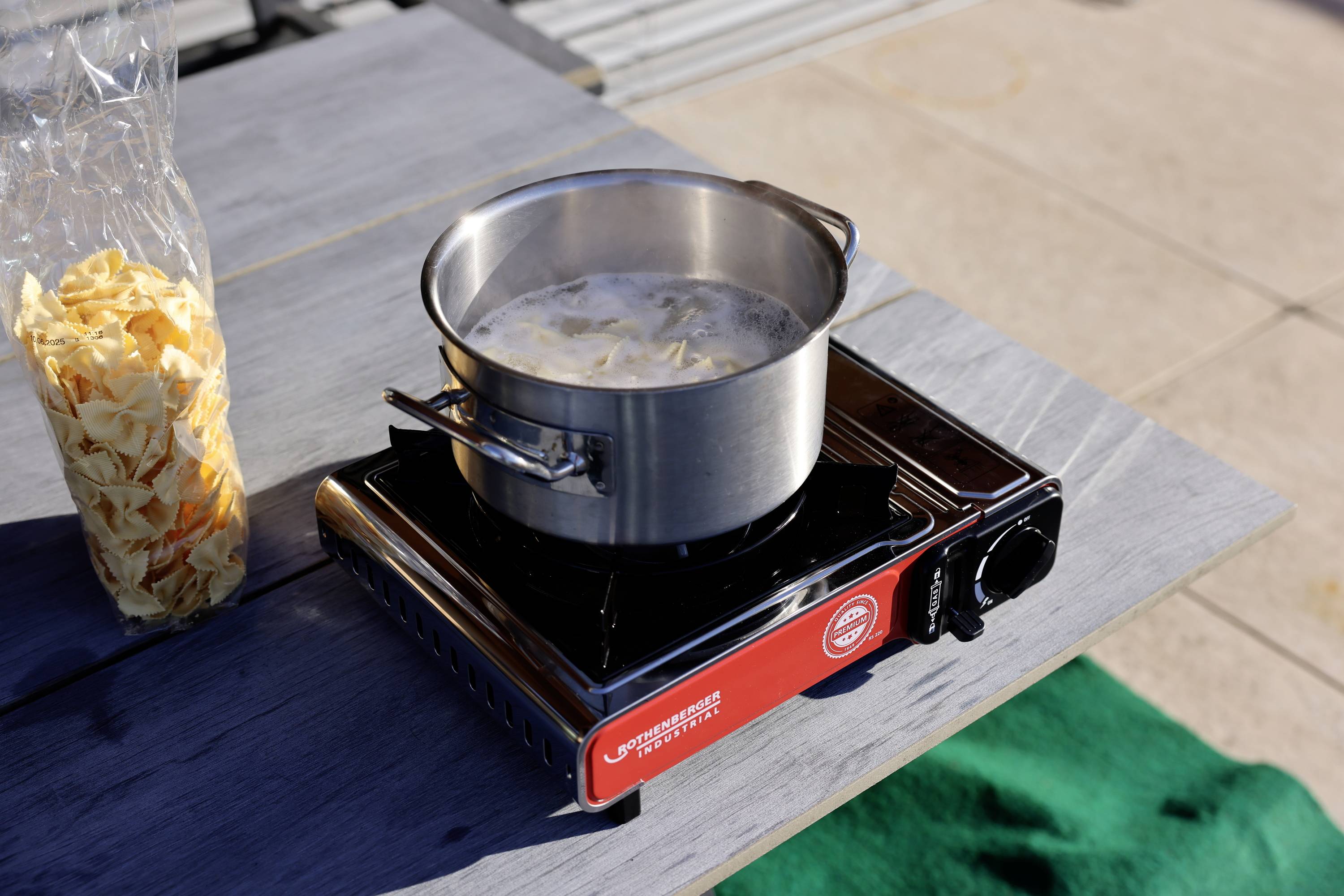 Een pan met kokend water en pasta staat op een draagbaar gasstel buiten. Naast de pan ligt een geopende verpakking pasta.