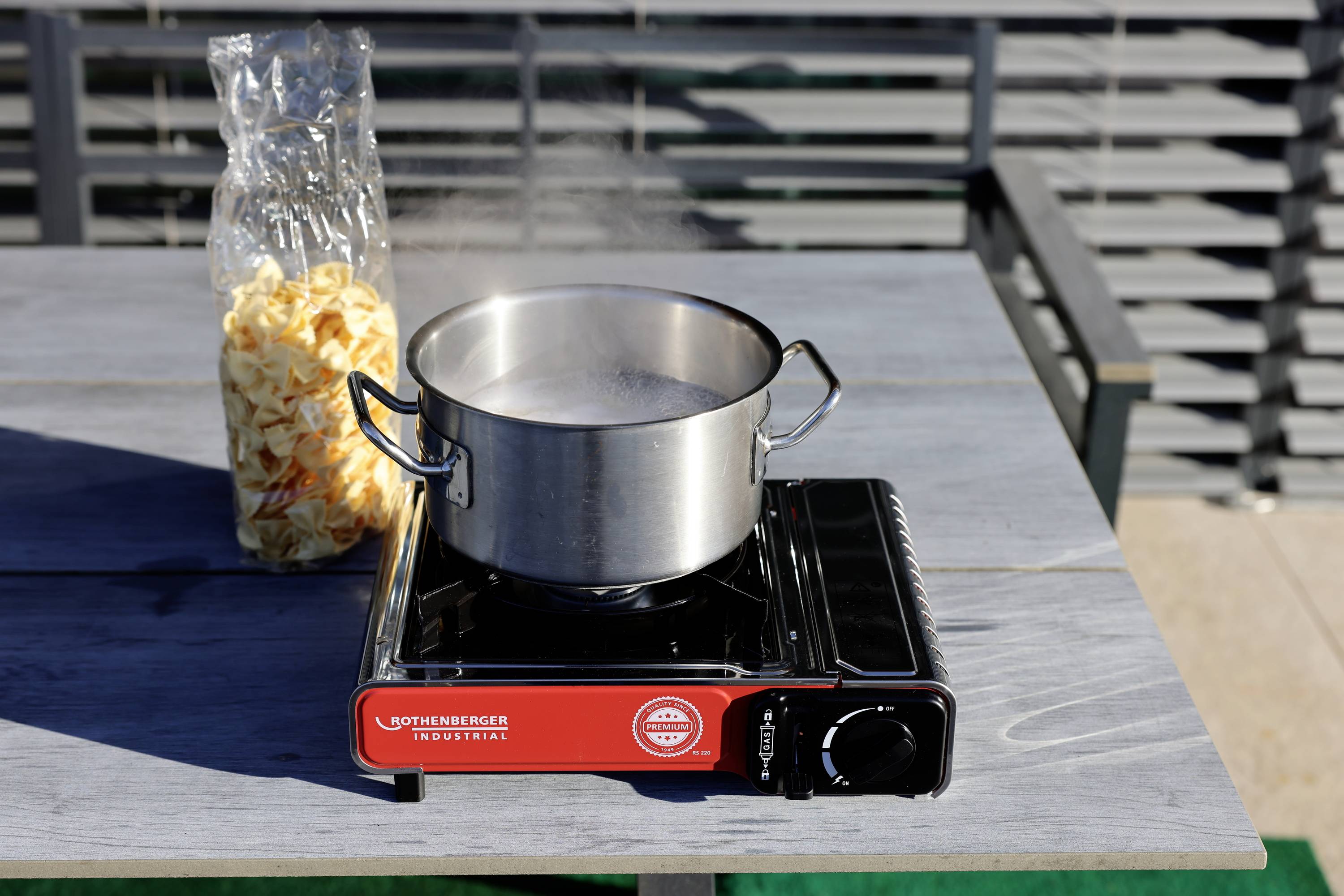 Een pot staat te koken op een draagbaar gasstel buiten. Ernaast staat een geopende zak met gedroogde pasta op tafel.