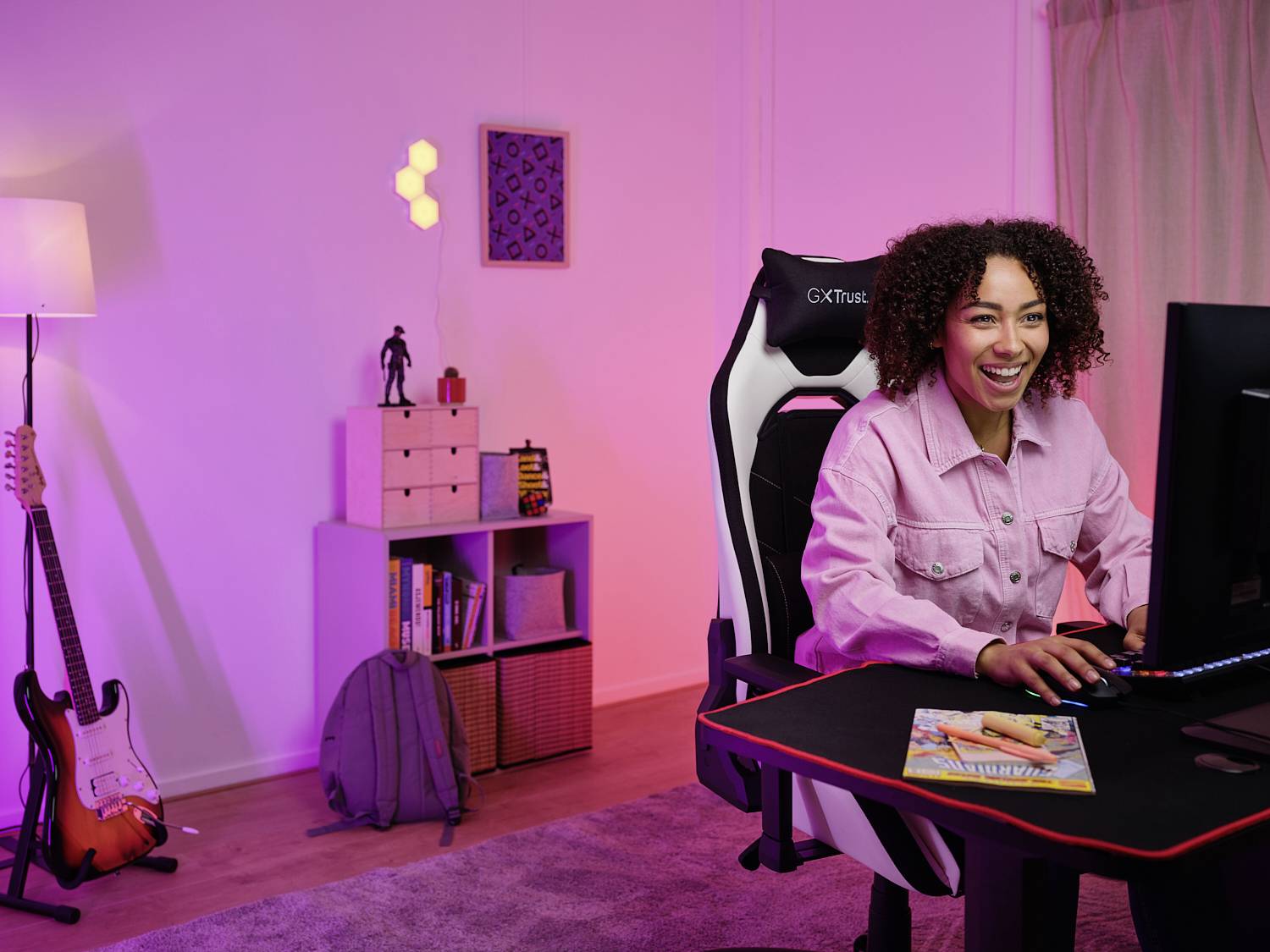 Een glimlachende persoon zit voor een computer in een gezellige kamer met gitaren en decoratie, omgeven door gekleurd licht.