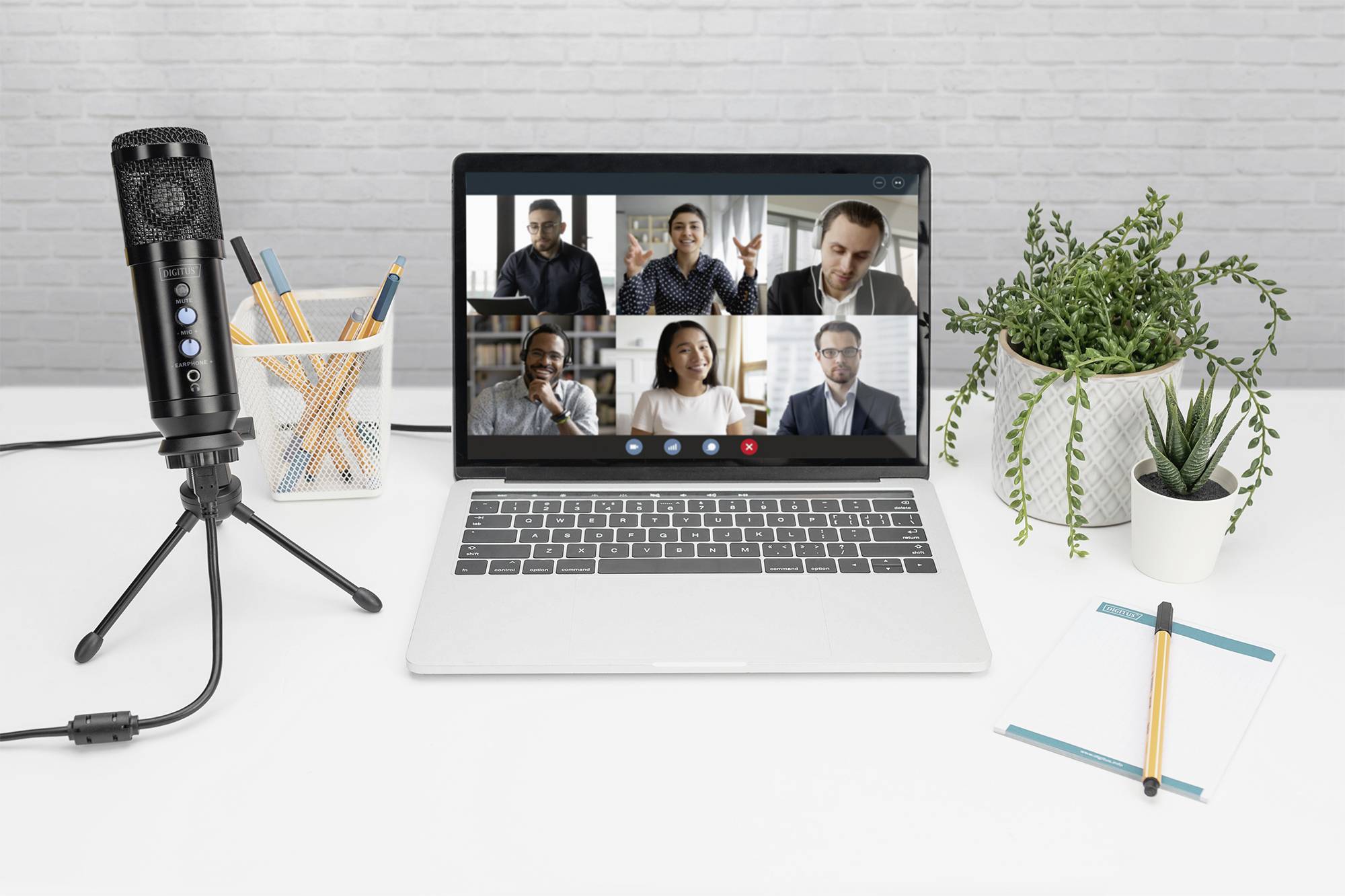 Laptop op een tafel toont videovergadering met zes personen. Links een microfoon, ernaast planten en schrijfbenodigdheden.
