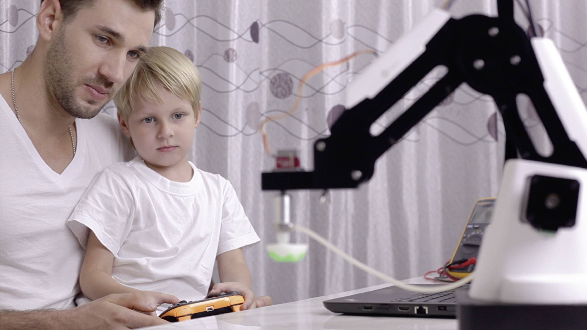 Een man en een kind zitten aan een tafel en kijken naar een laptop, terwijl een mechanische arm op de voorgrond zichtbaar is.