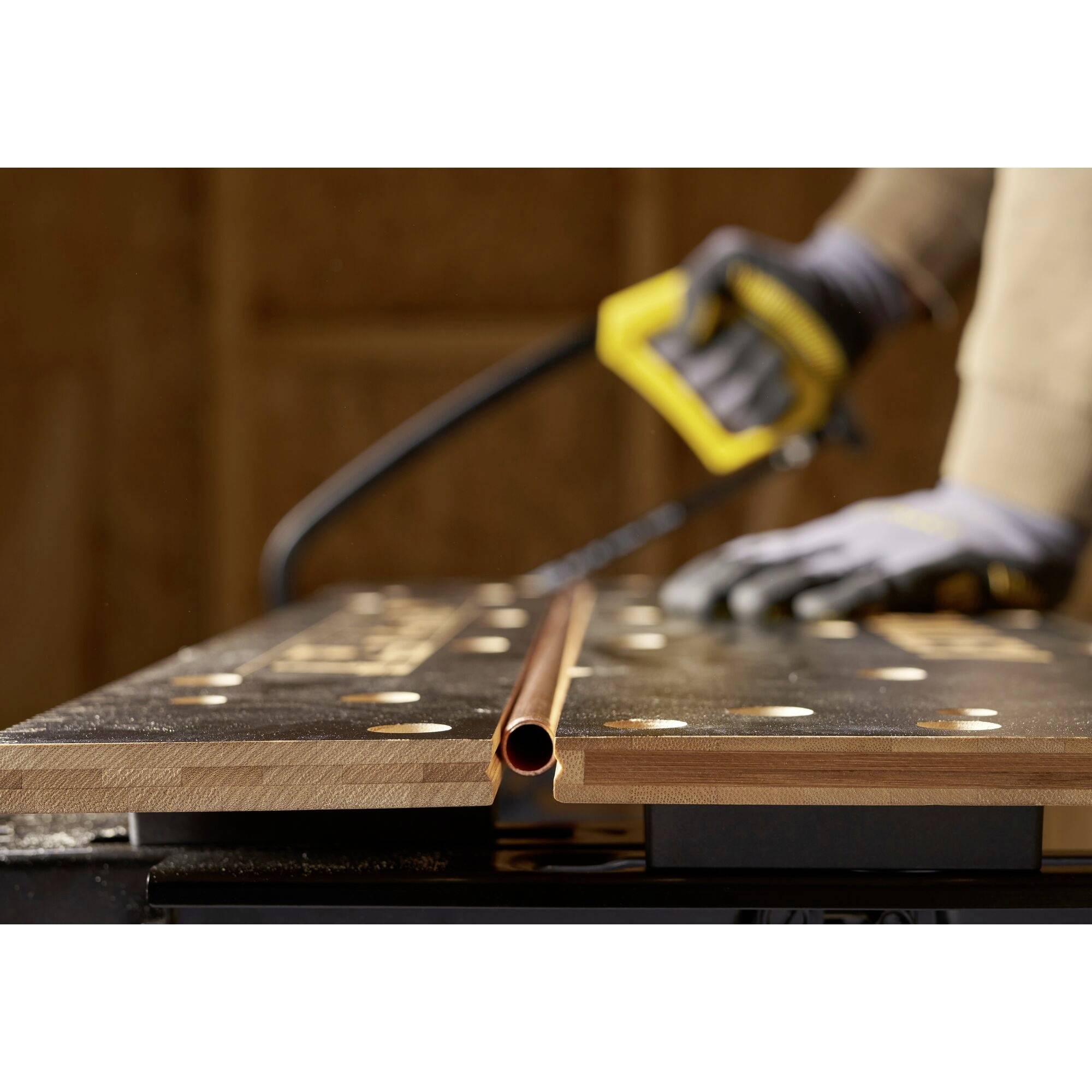 Een persoon zaagt een koperen buis op een werkbank met een handzaag. De focus ligt op de buis en de zaag in de hand.