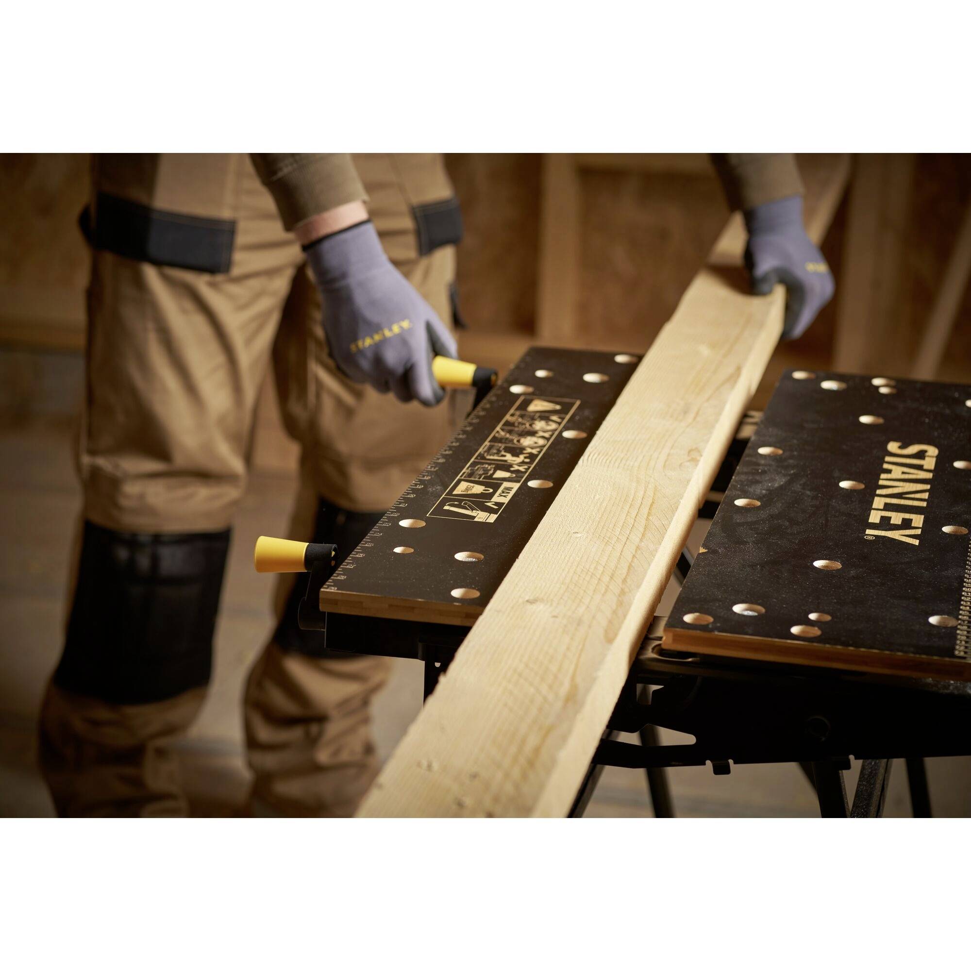 Een persoon zaagt een lang houten plank op een werkbank. Ze draagt werkkleding en handschoenen. Op de achtergrond is een werkplaats.