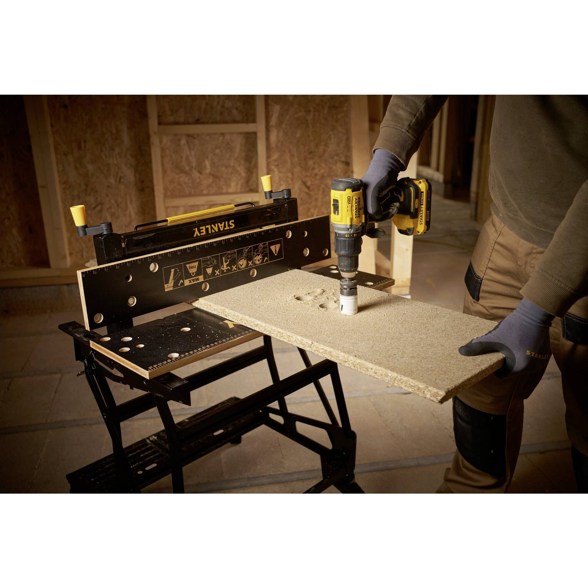 Een persoon boort met een gele boormachine een groot gat in een spaanplaat die op een zwarte werktafel ligt.