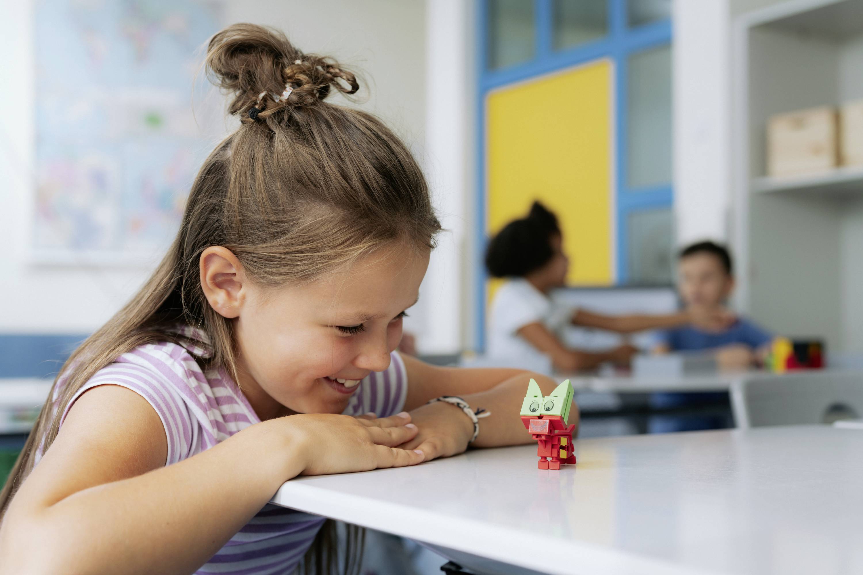 Een meisje lacht naar een speelgoed dat op een tafel in een klaslokaal staat. Op de achtergrond zijn twee kinderen onscherp.