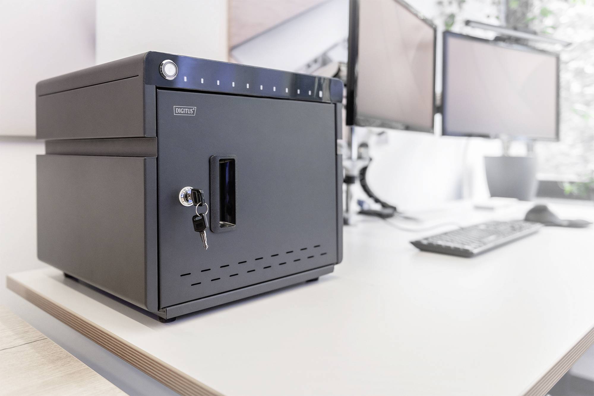 Een zwarte kluis met sleutel ligt op een lichte bureau; op de achtergrond zijn twee computermonitoren zichtbaar.