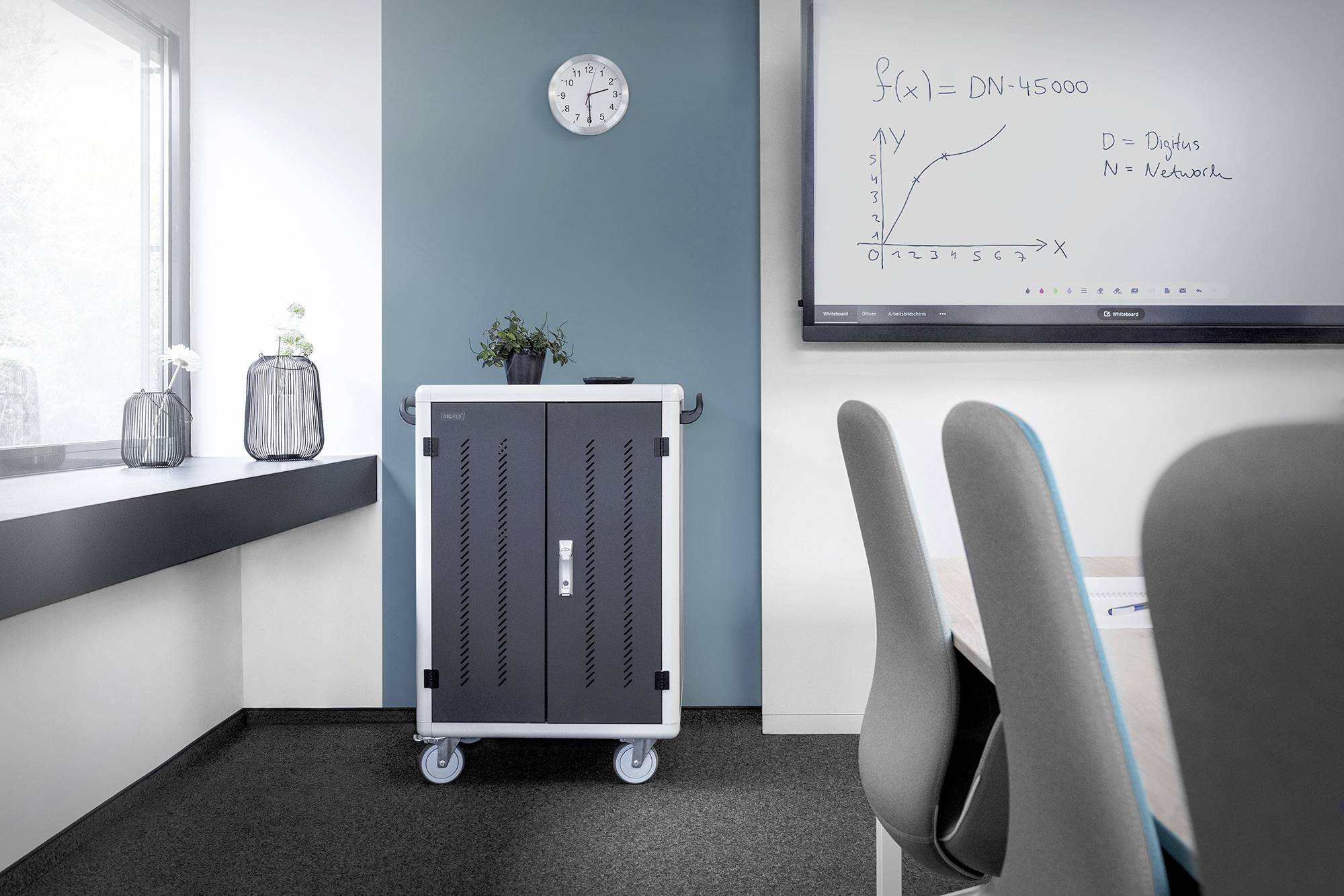 Een vergaderzaal met een tafel en stoelen, een verrijdbare kast, een whiteboard met een getekende functie en een klok aan de muur.