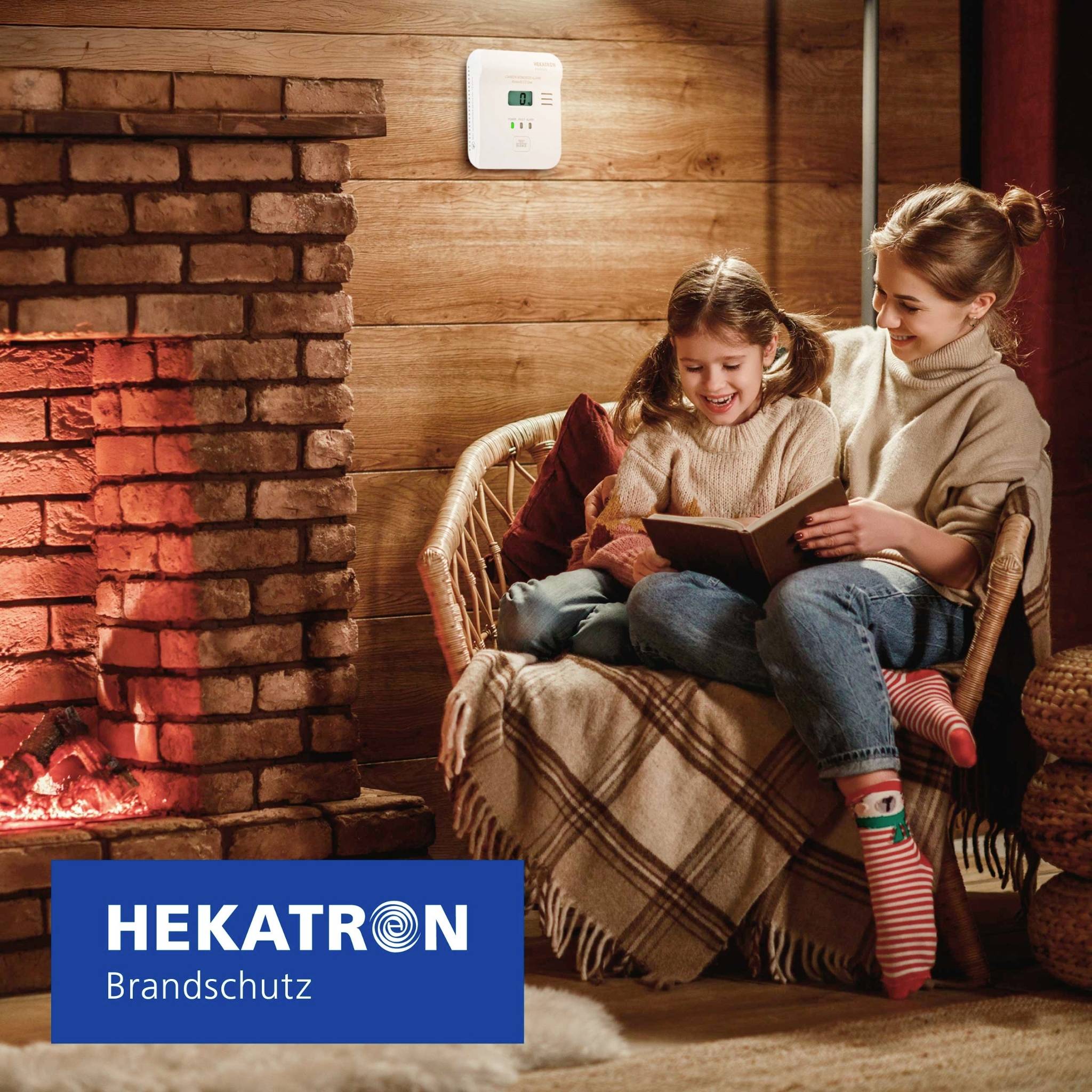 Een vrouw en een kind zitten bij een brandende open haard, samen te lezen. Een brandveiligheidsapparaat is aan de muur bevestigd. Het merkenlogo, 'HEKATRON Brandschutz', is zichtbaar.