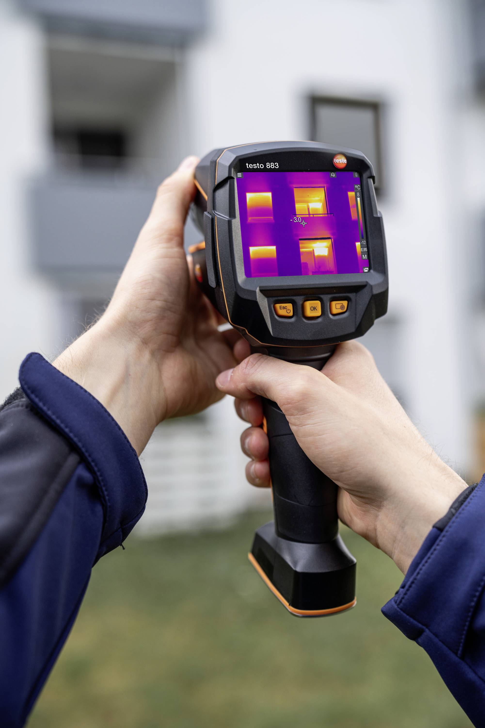 Een persoon houdt een warmtebeeldcamera vast die de temperatuurverdeling op een gebouwoppervlak weergeeft. Gekleurde gebieden tonen temperatuurverschillen.