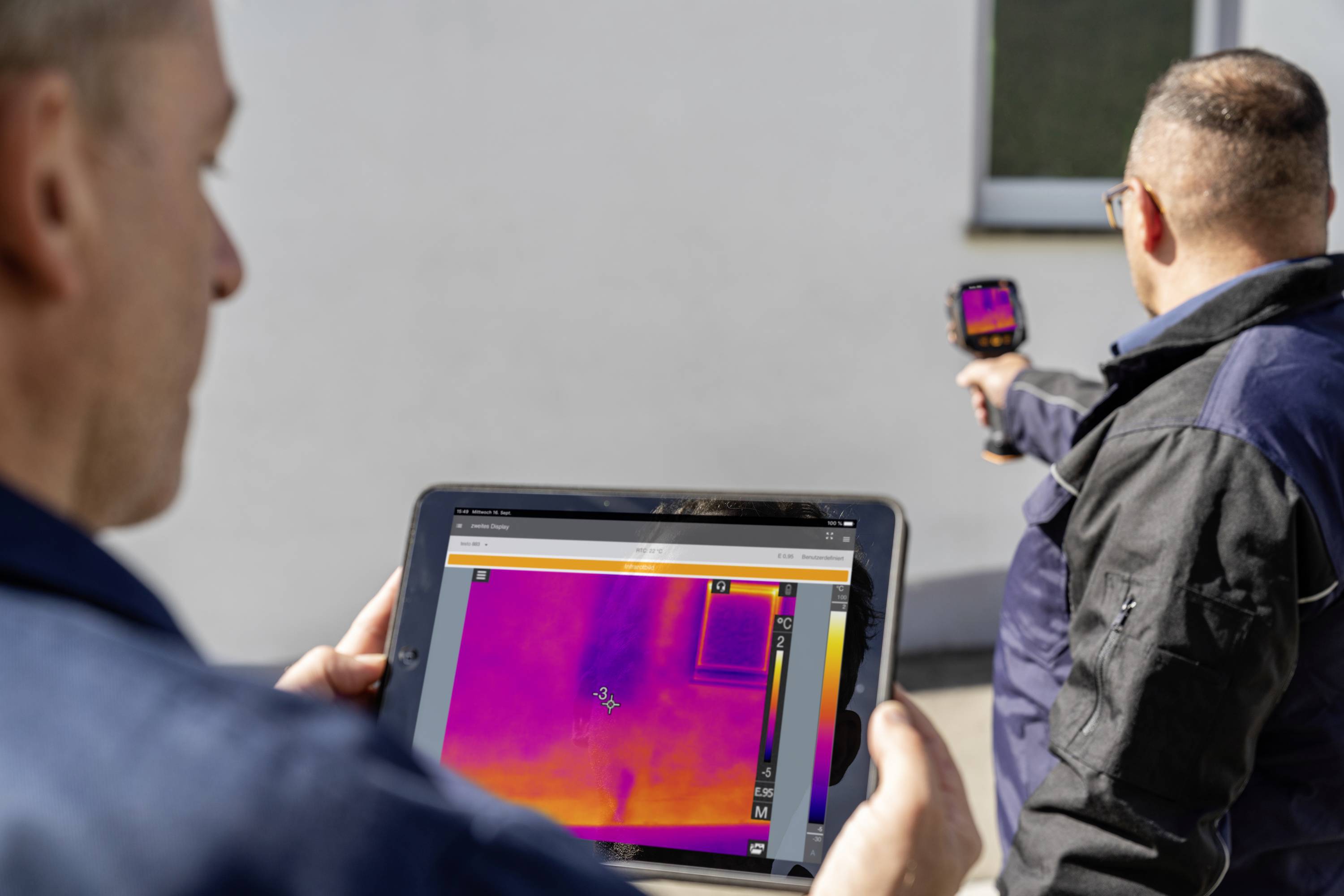 Twee mannen voeren een warmtebeeldonderzoek uit aan een muur, waarbij een van hen een tablet vasthoudt met een kleurenscherm en de ander een camera.