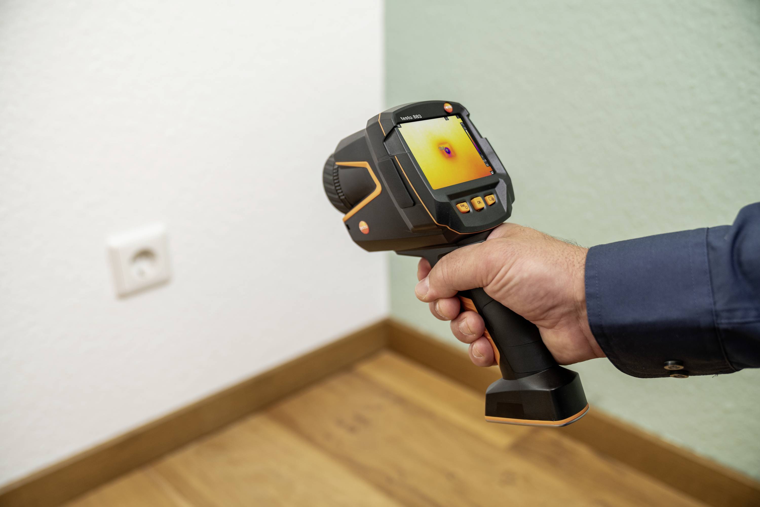 Een persoon houdt een warmtebeeldcamera en richt deze op een muur in de kamer naast een stopcontact. Op het scherm verschijnt een warmtebeeld.