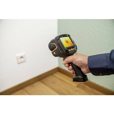 Een persoon houdt een warmtebeeldcamera en richt deze op een muur in de kamer naast een stopcontact. Op het scherm verschijnt een warmtebeeld.