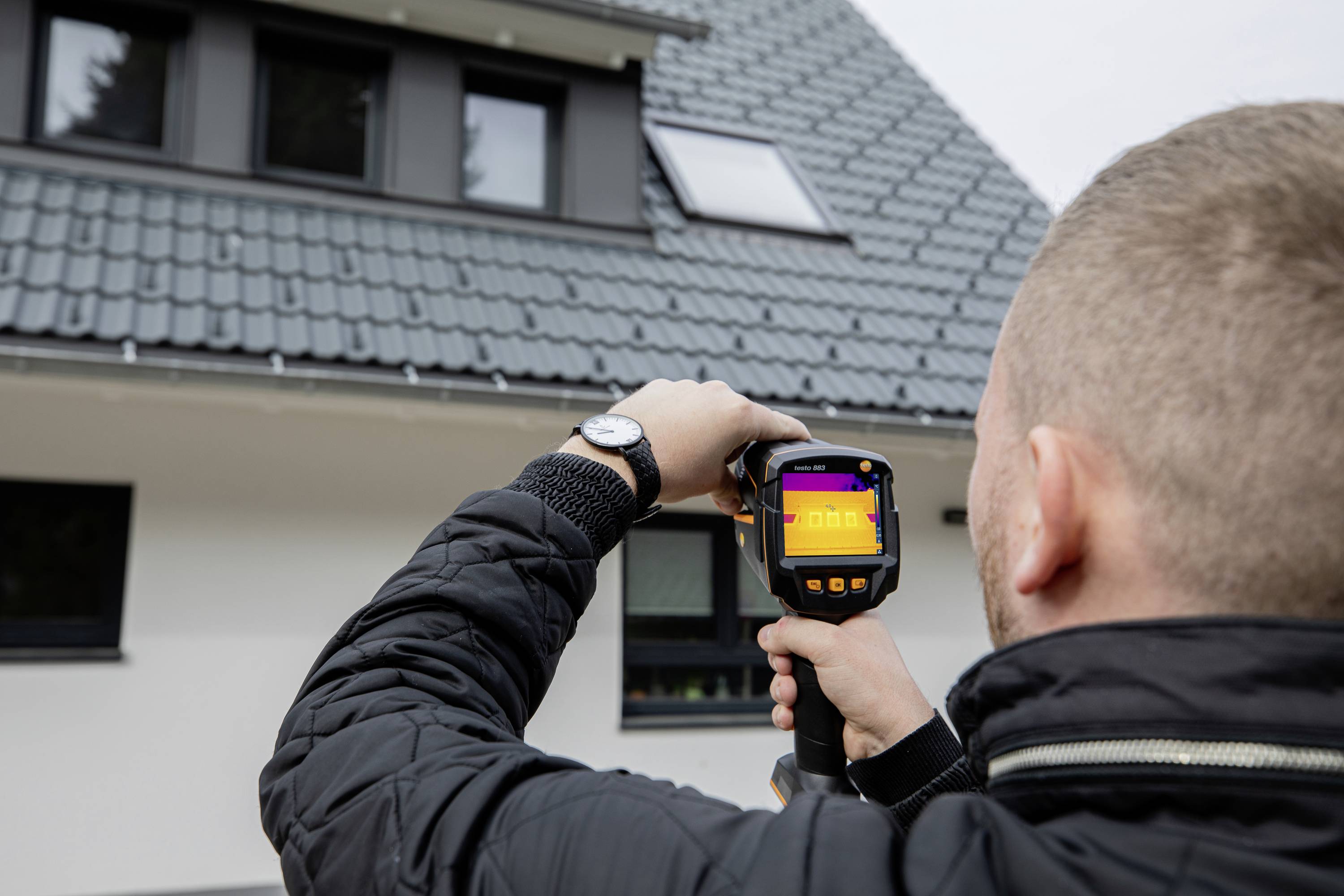 Een persoon houdt een warmtebeeldcamera voor een huis en controleert de dakisolatie.