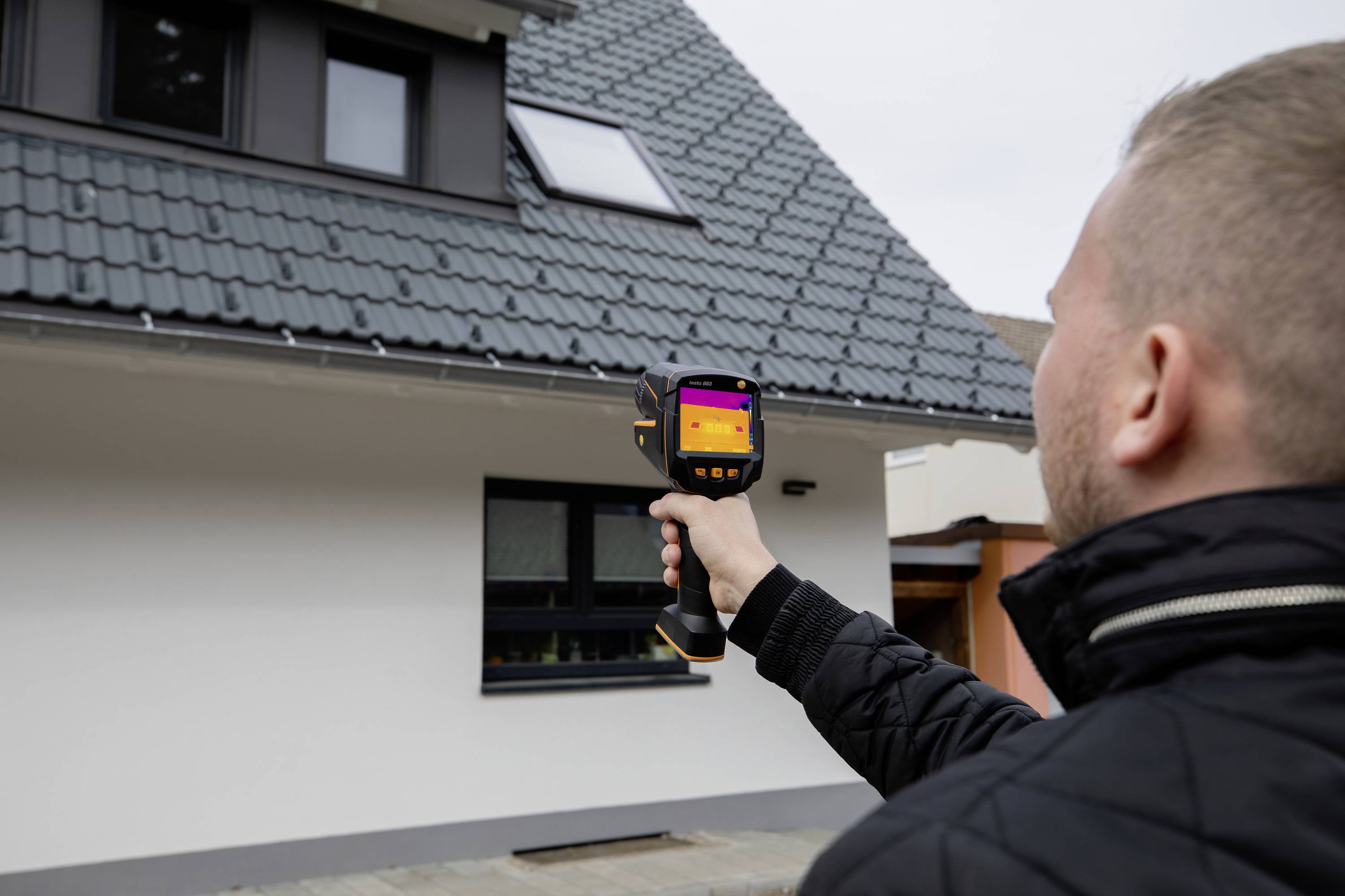 Een man gebruikt een warmtebeeldcamera om een huis te inspecteren. De camera toont temperatuurverschillen op de gevel.