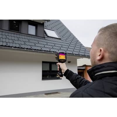 Een man gebruikt een warmtebeeldcamera om een huis te inspecteren. De camera toont temperatuurverschillen op de gevel.