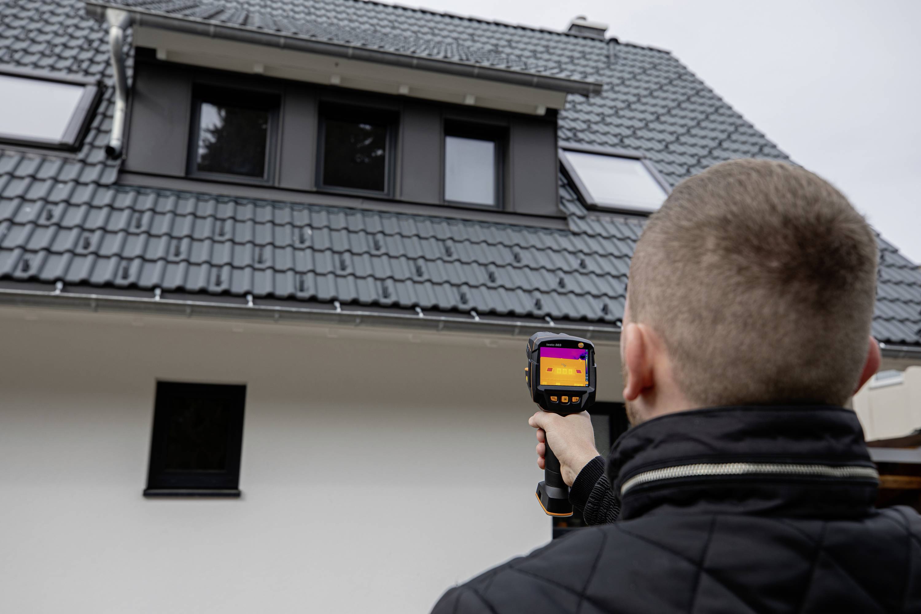 Een persoon richt een warmtebeeldcamera op het dak van een huis. De camera toont een warmtebeeld ter controle van de isolatie.