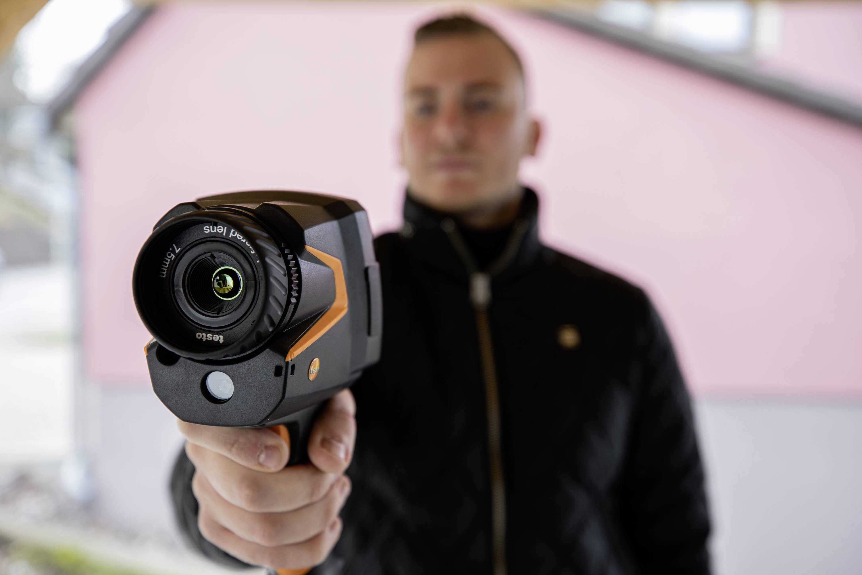 Een persoon houdt een warmtebeeldcamera die op de toeschouwer is gericht. Op de achtergrond is een onscherpe roze muur zichtbaar.