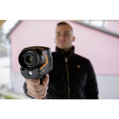 Een persoon houdt een warmtebeeldcamera die op de toeschouwer is gericht. Op de achtergrond is een onscherpe roze muur zichtbaar.