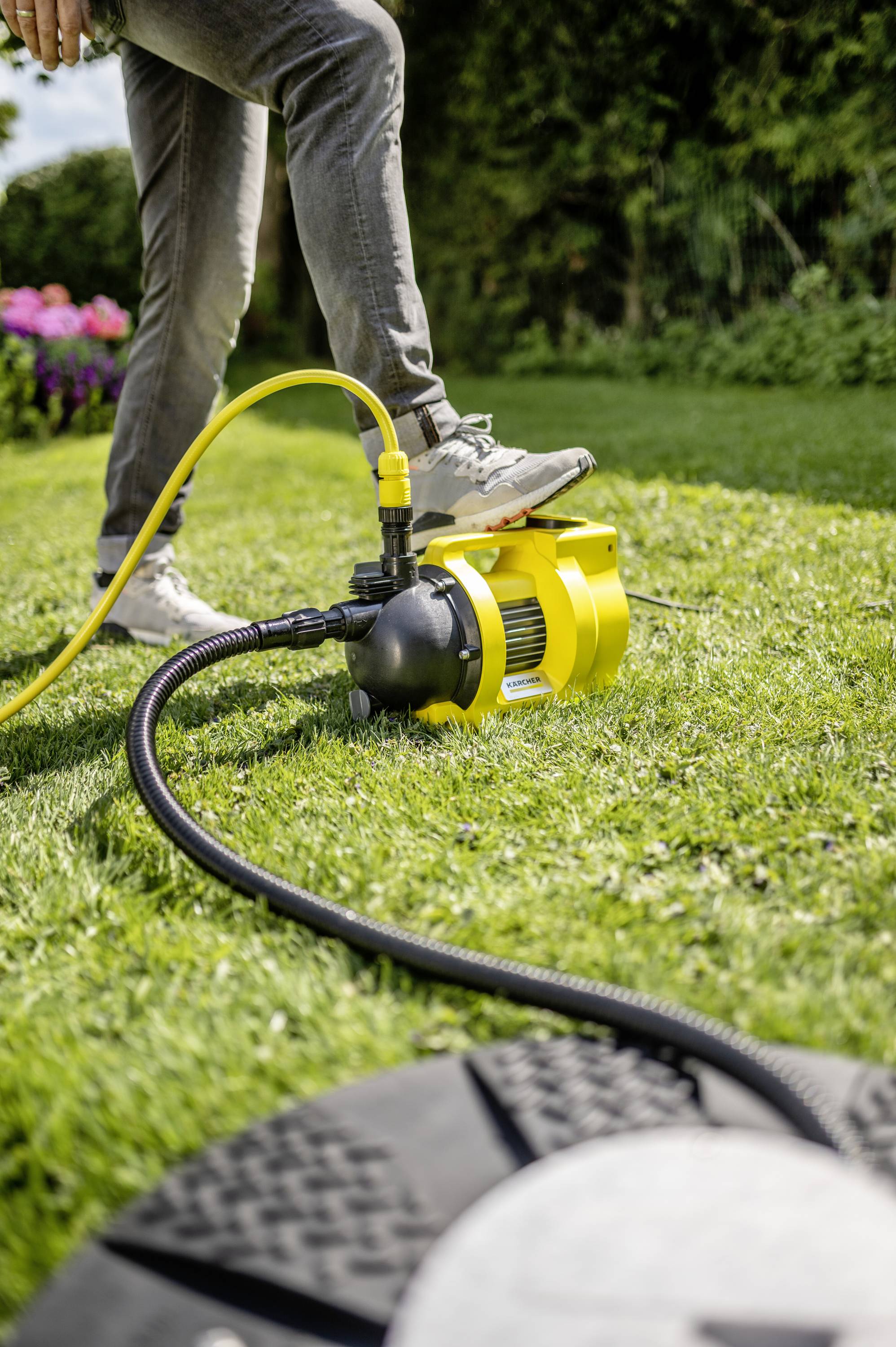 Een persoon gebruikt een gele waterpomp om het gazon in een tuin te besproeien. De omgeving is groen en bloemrijk.