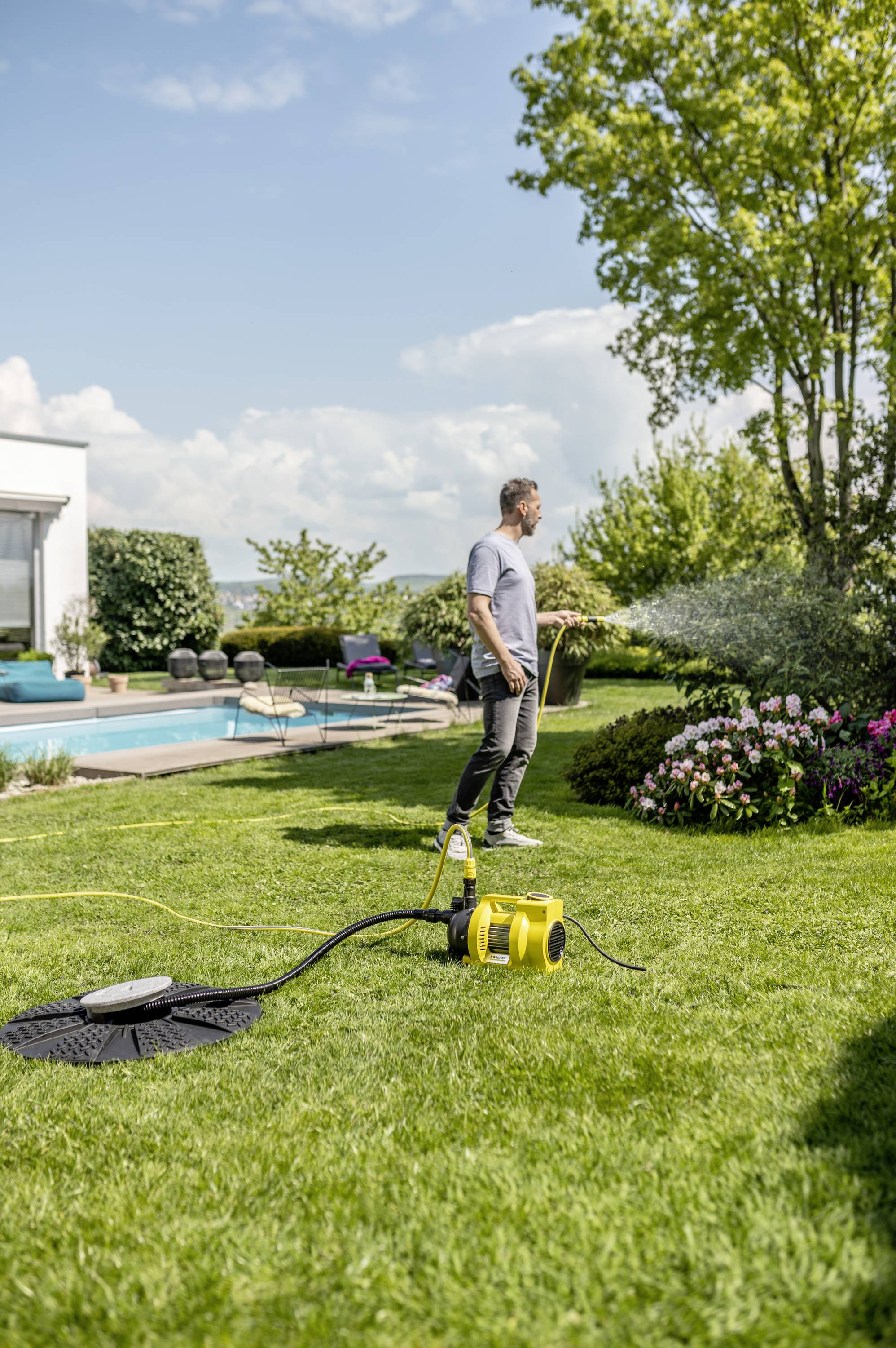 Een man besproeit zijn gazon in de tuin met een automatische sproeier. Op de achtergrond is een modern huis met een zwembad te zien.