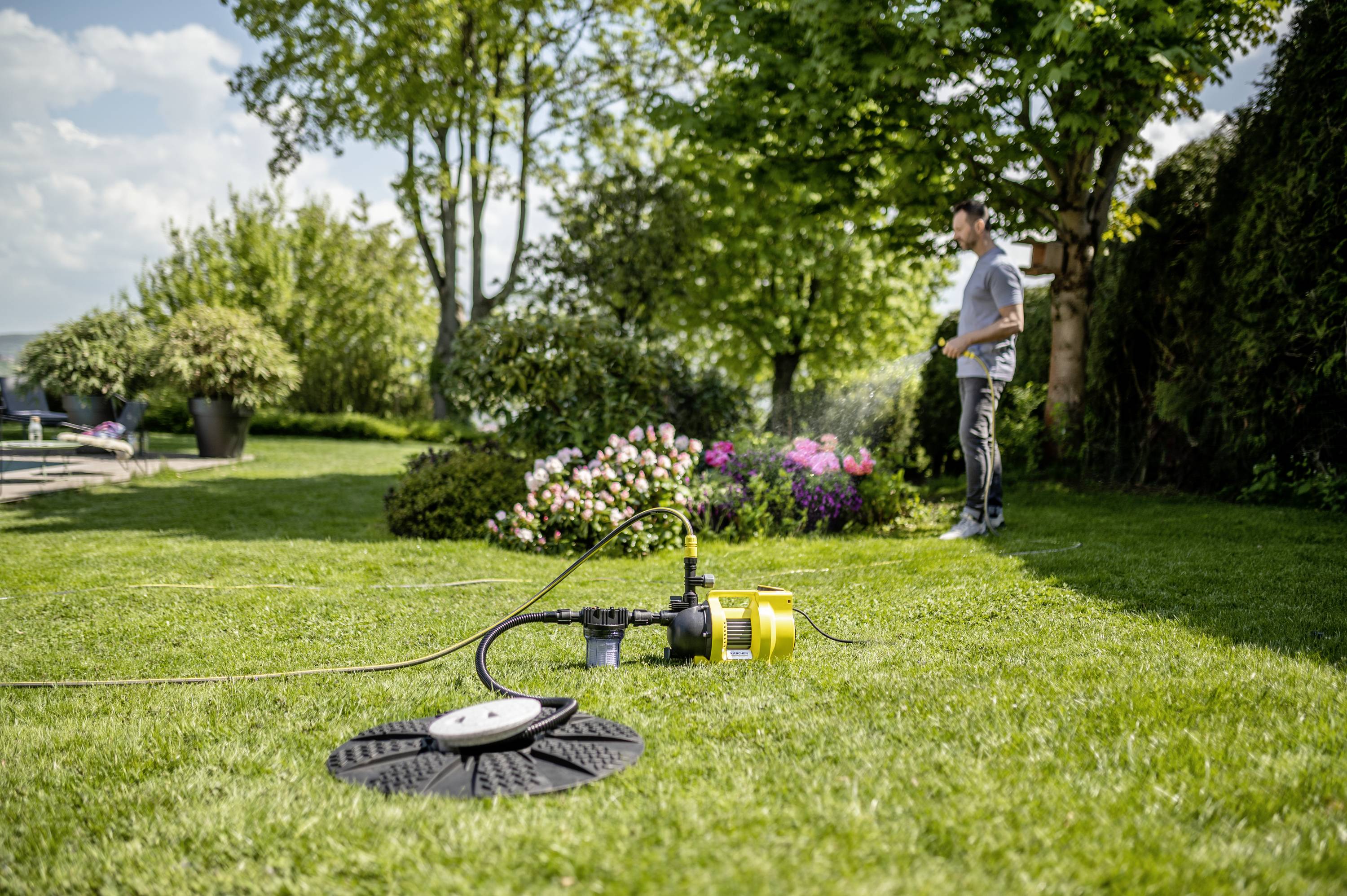 Een man staat in de tuin naast een groene weide, waarop een geel irrigatieapparaat water verspreidt. Op de achtergrond bloeien struiken.