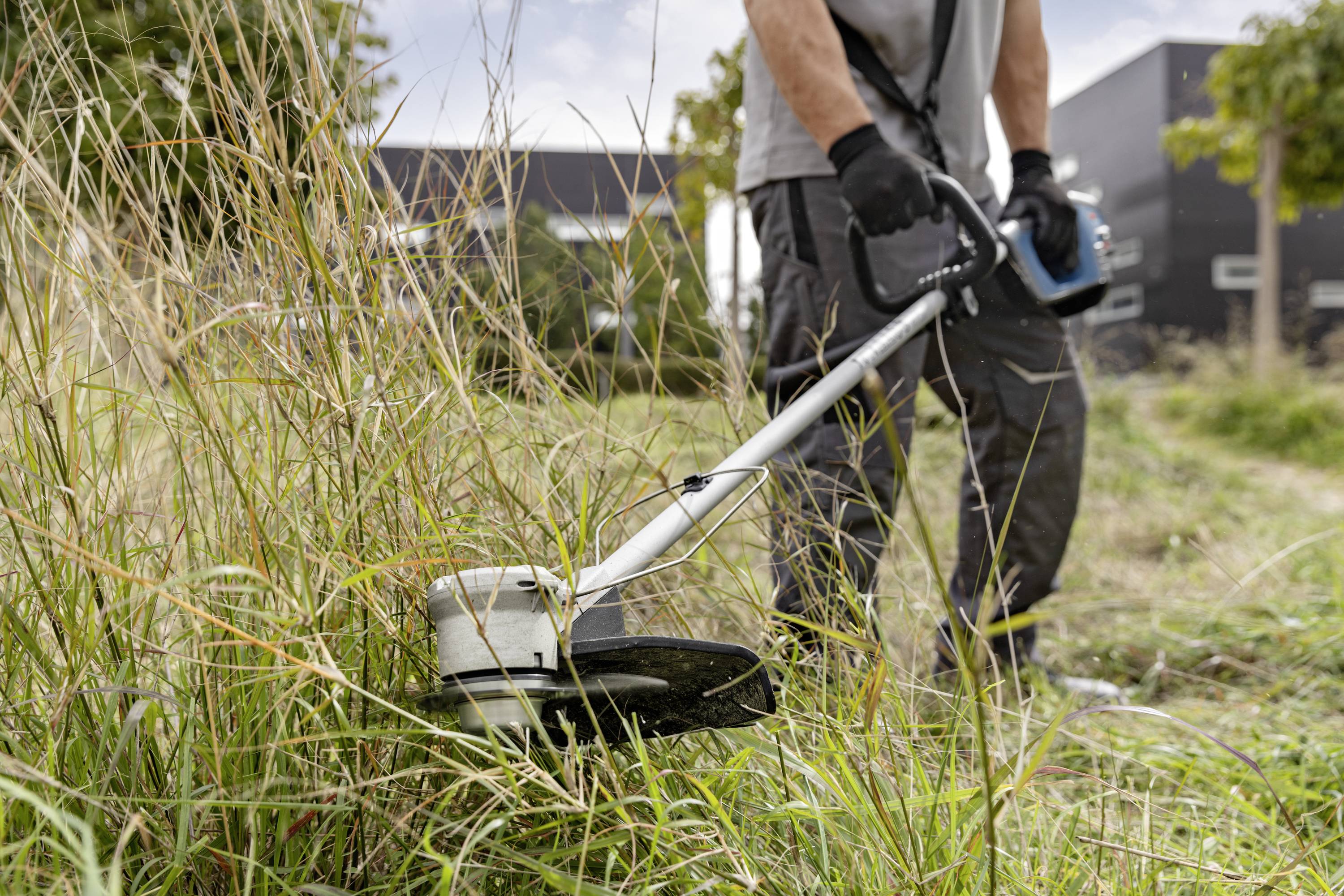 Een persoon maait hoog gras met een grastrimmer op een weide. Op de achtergrond zijn bomen en gebouwen zichtbaar.