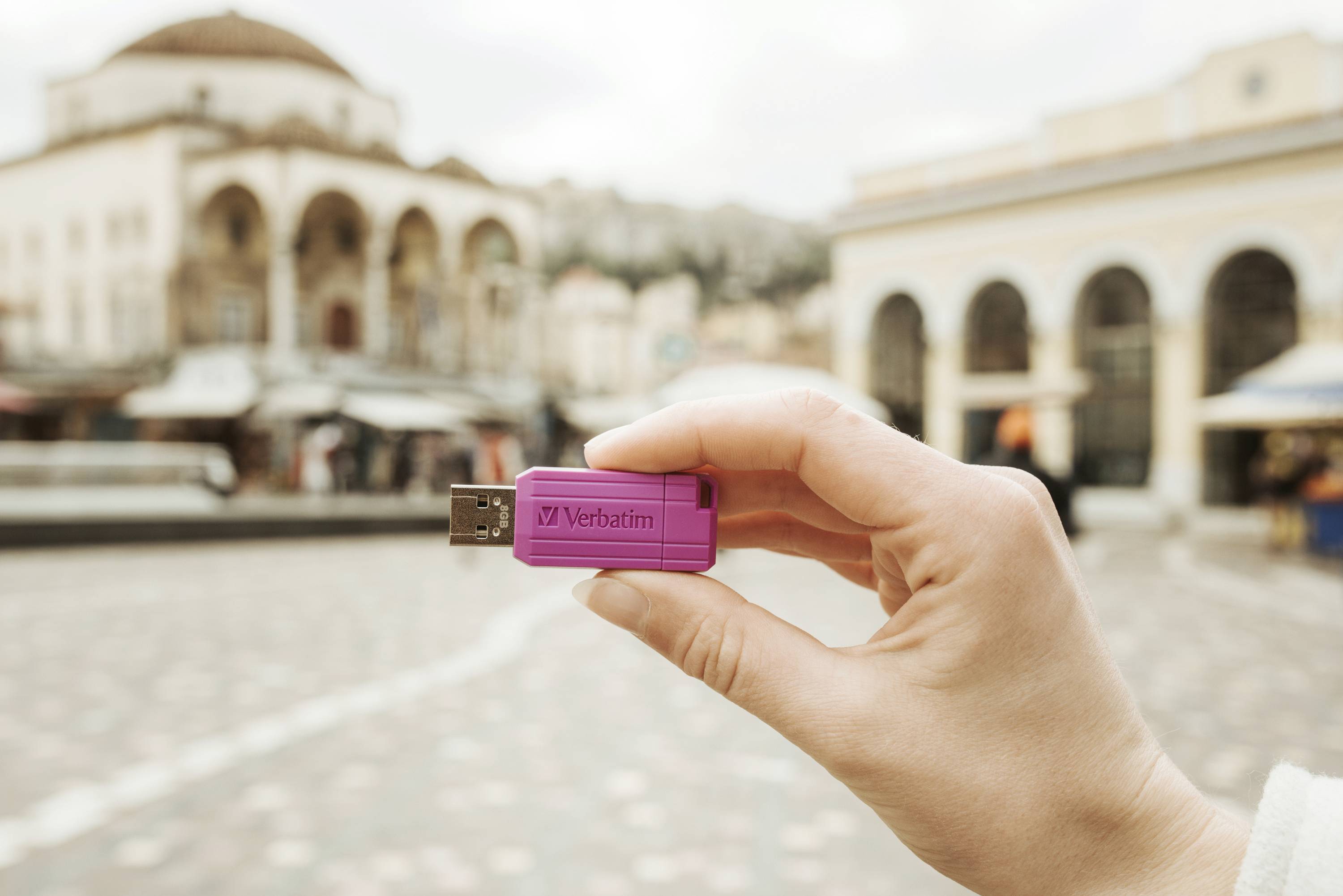 Een hand houdt een paarse USB-stick met het opschrift 'Verbatim' vast voor een onscherpe achtergrond met gebouwen.