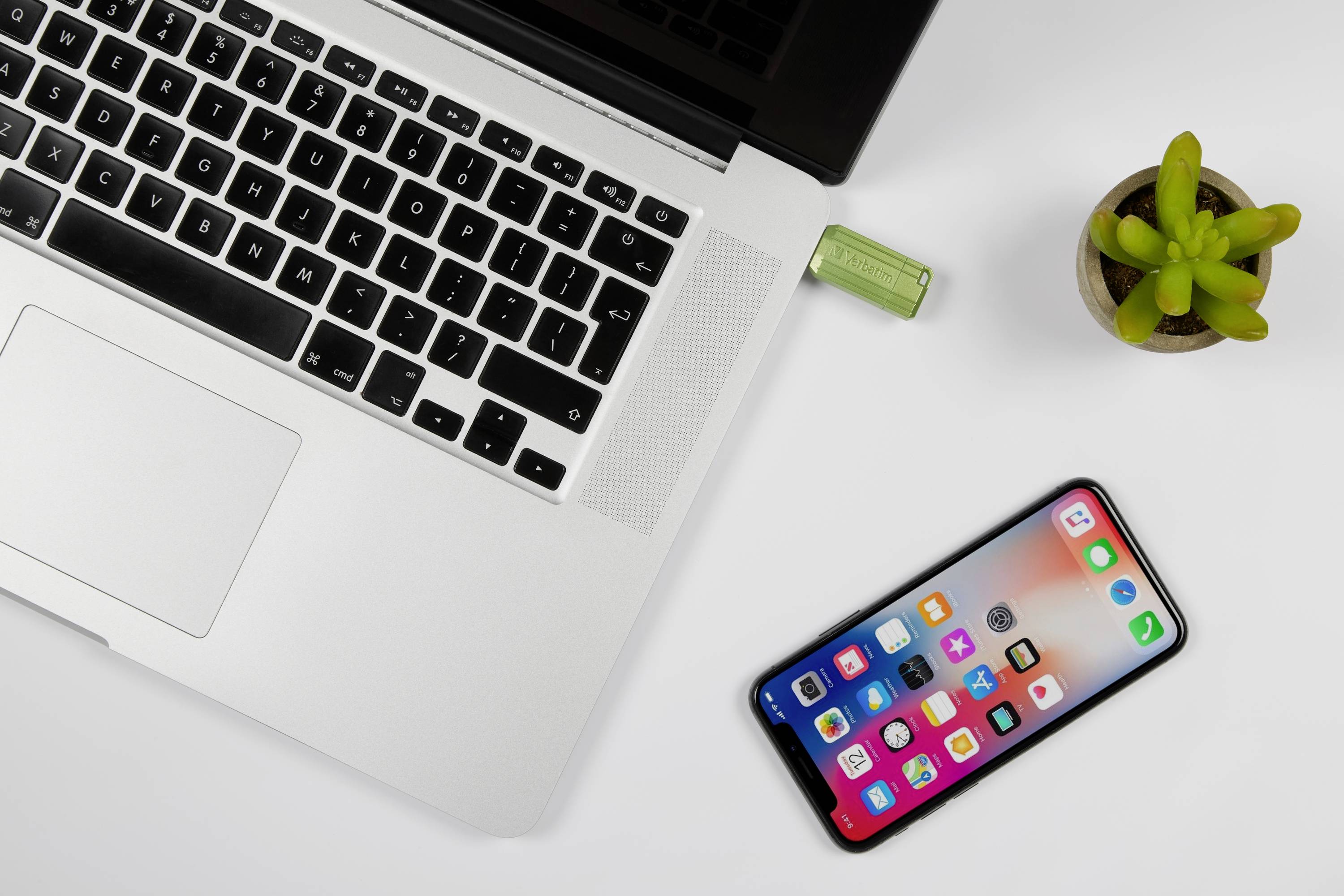 Op een bureau liggen een opengeslagen laptop, een ingestoken USB-stick, een smartphone met apps en een kleine plant.