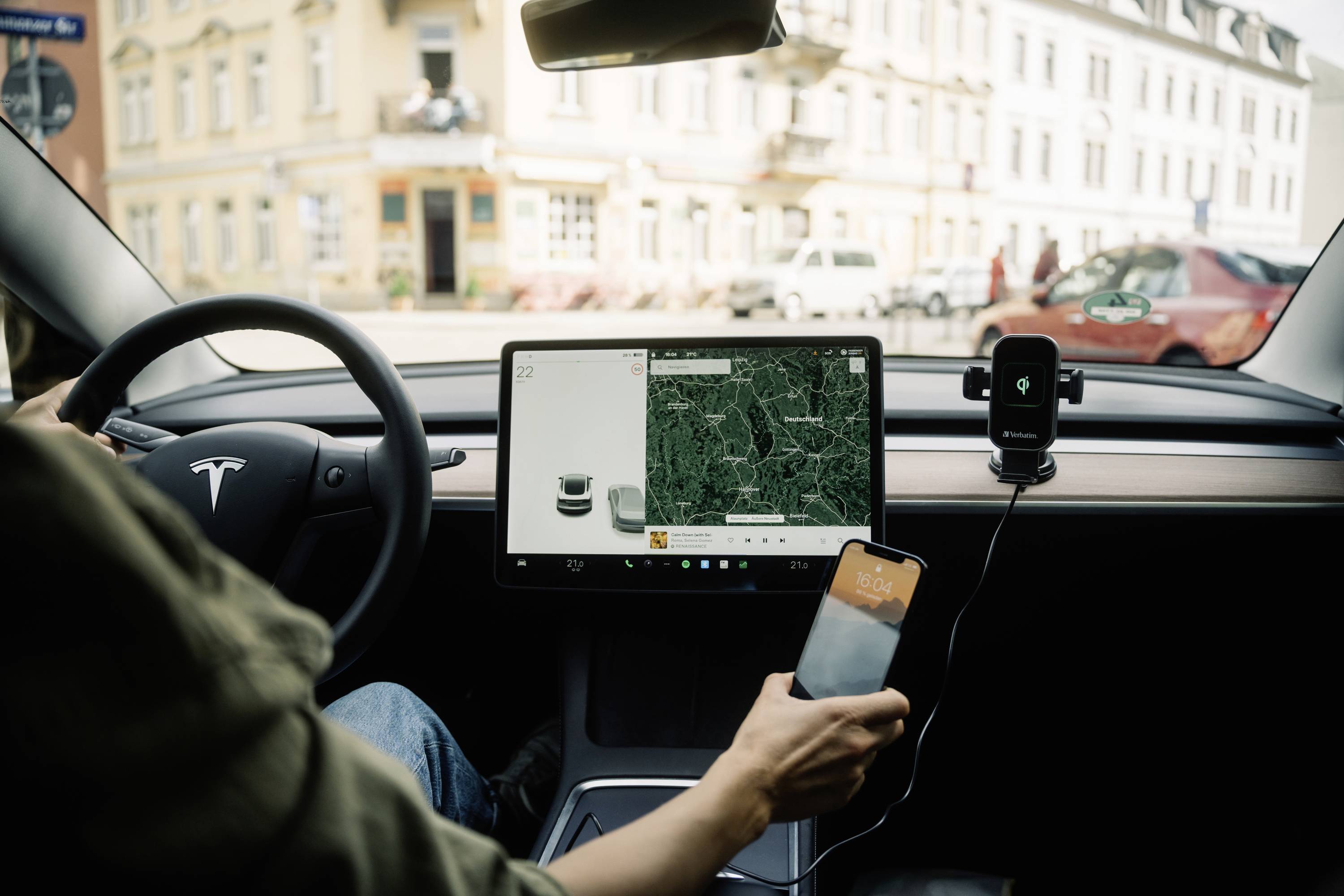 Persoon rijdt auto met groot touchscreen-display, toont kaartapplicatie. Buitenaanzicht van een stad met gebouwen en geparkeerde auto's.