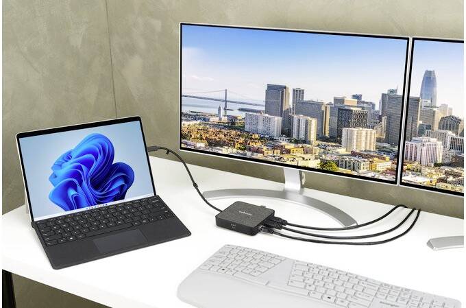 Een bureau met een laptop die via kabels is aangesloten op een dockingstation. Het dockingstation is verbonden met twee monitors die een stadsaanzicht laten zien.