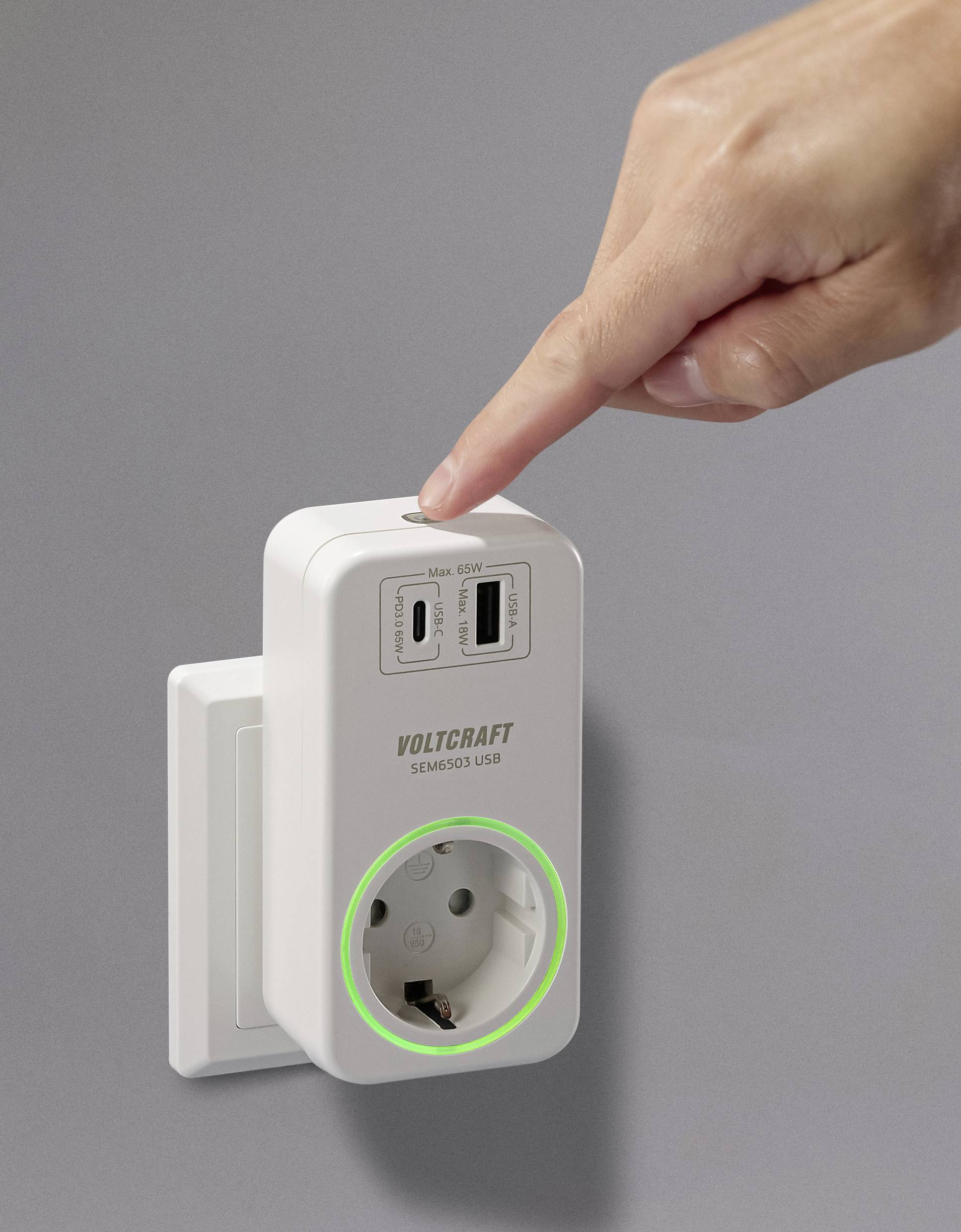Een hand wijst naar een witte stekkerdoos-adapter met USB-poorten en een groene verlichte ringaanduiding, die aan een muur is bevestigd.