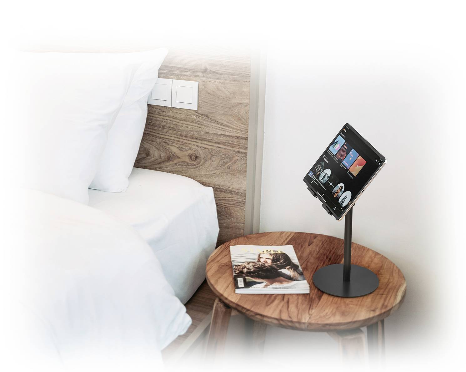 Een tablet op een nachtkastje naast een bed toont een app met entertainmentaanbiedingen. Daaronder ligt een tijdschrift.