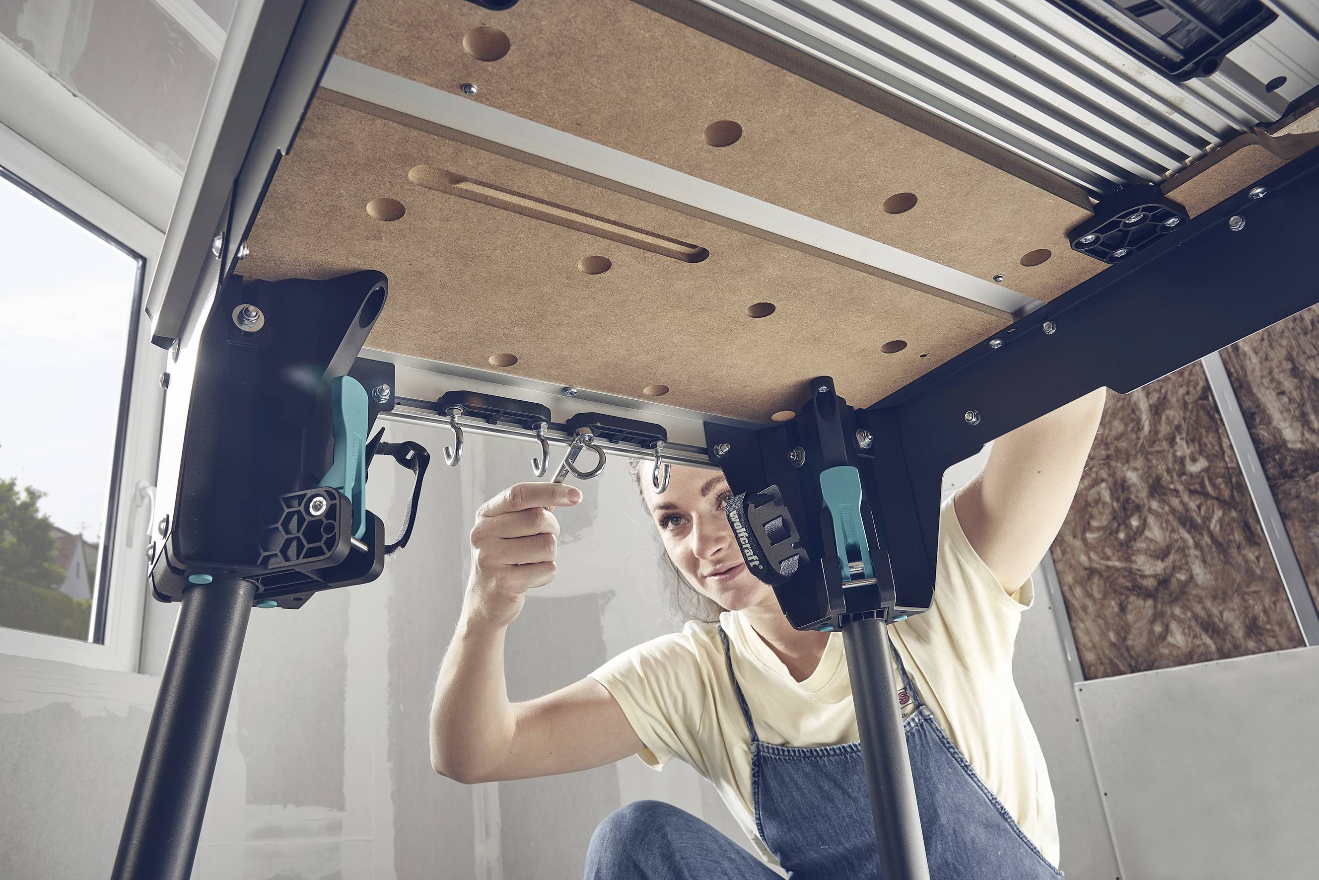 Een persoon monteert een tafel met een zwart frame en een houten blad in een lichte ruimte. Daglicht valt door het raam naar binnen.