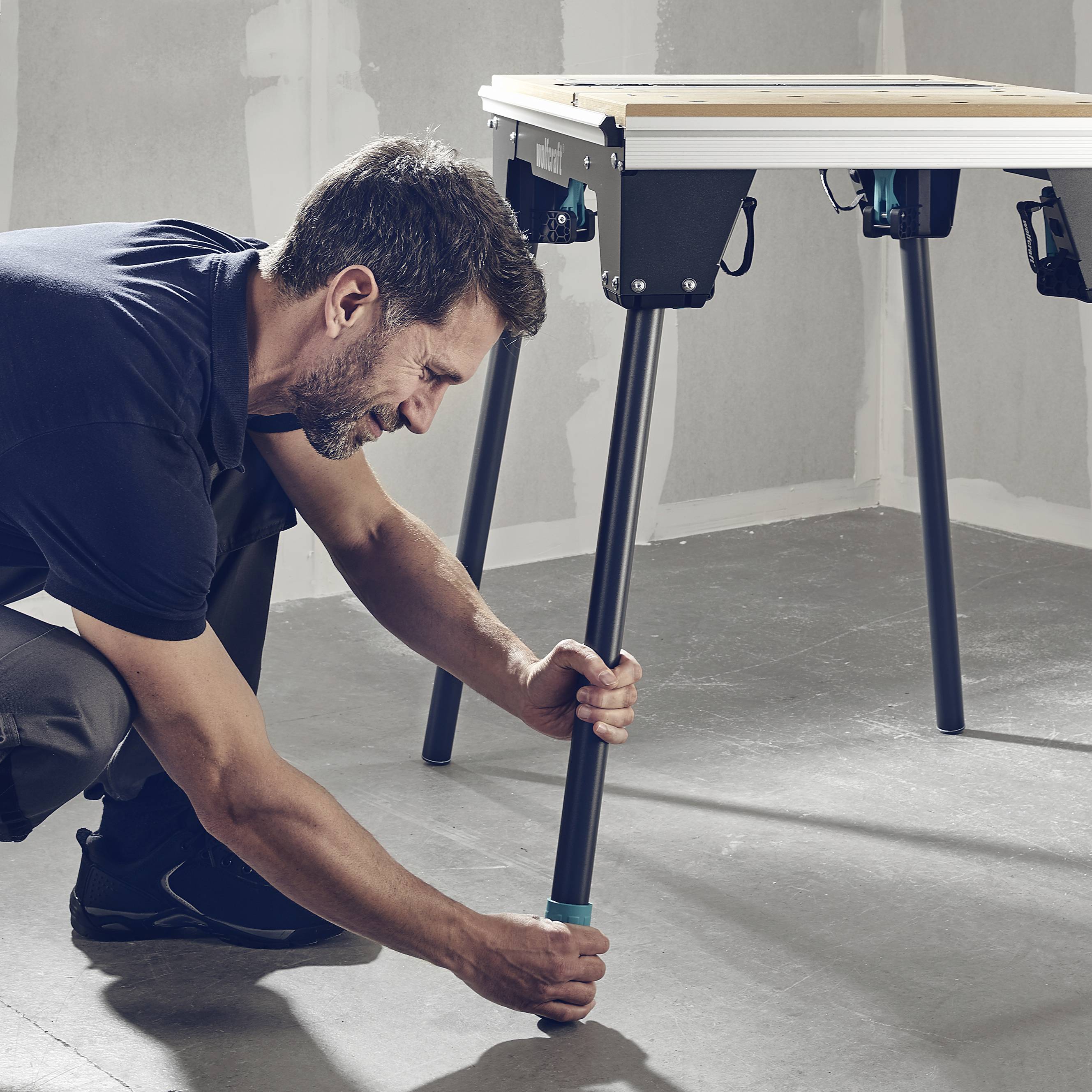 Een man stelt de hoogte van een tafelpoot bij in een werkplaats. De tafel heeft een lichte oppervlakte en staat op een grijze vloer.