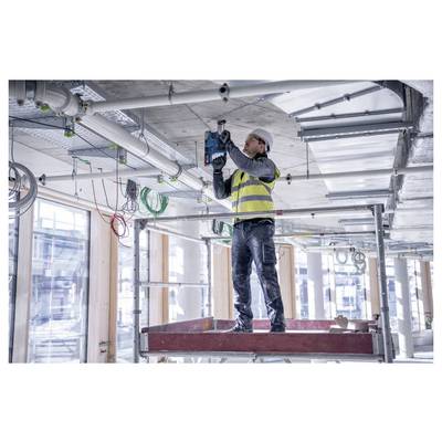 Een bouwvakker met een veiligheidshelm en vest gebruikt een elektrisch gereedschap om in het plafond van een grote, onafgewerkte binnenruimte te boren.