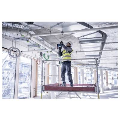 Een bouwvakker in een veiligheidshesje en helm gebruikt een boormachine om apparatuur te installeren aan het plafond van een modern gebouw in aanbouw.