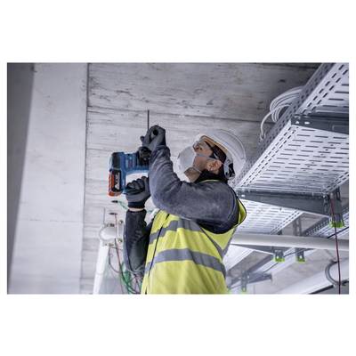 Een bouwvakker met een veiligheidshelm en een reflecterend veiligheidshesje boort met een elektrische boormachine in een plafond, omringd door elektriciteitskabels en leidingen.