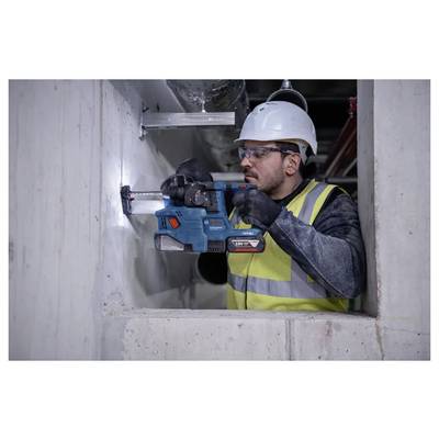 Een bouwvakker met een veiligheidshelm en een reflecterend veiligheidshesje boort met een elektrische boormachine in een betonnen muur binnen in een gebouw in aanbouw.