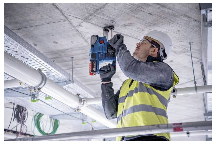 Een werknemer in een felgeel veiligheidshesje en helm gebruikt een elektrisch gereedschap om in een betonnen plafond te boren op een bouwplaats.