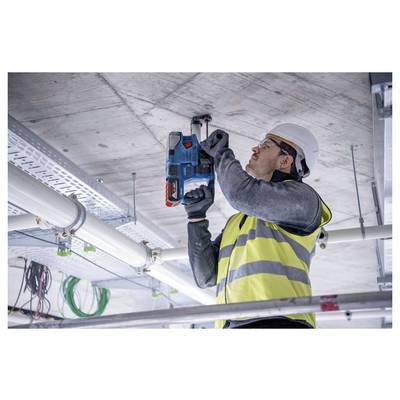 Een werknemer in een felgeel veiligheidshesje en helm gebruikt een elektrisch gereedschap om in een betonnen plafond te boren op een bouwplaats.