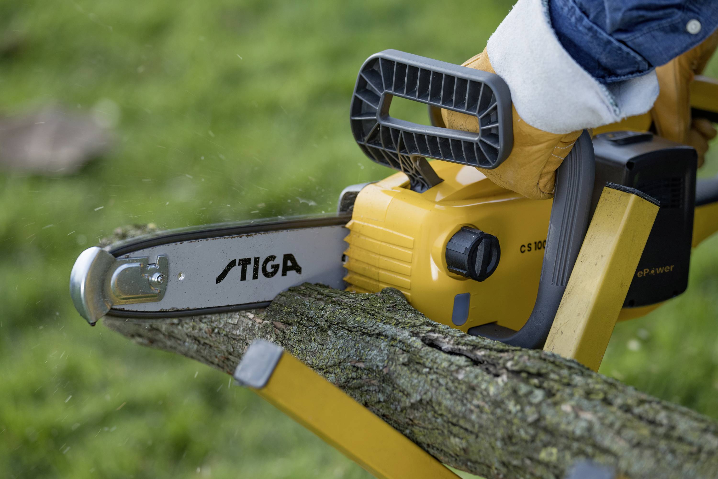 Een hand met handschoen houdt een kettingzaag vast die een tak aan het zagen is. De kettingzaag is geel met het opschrift 'STIGA'.