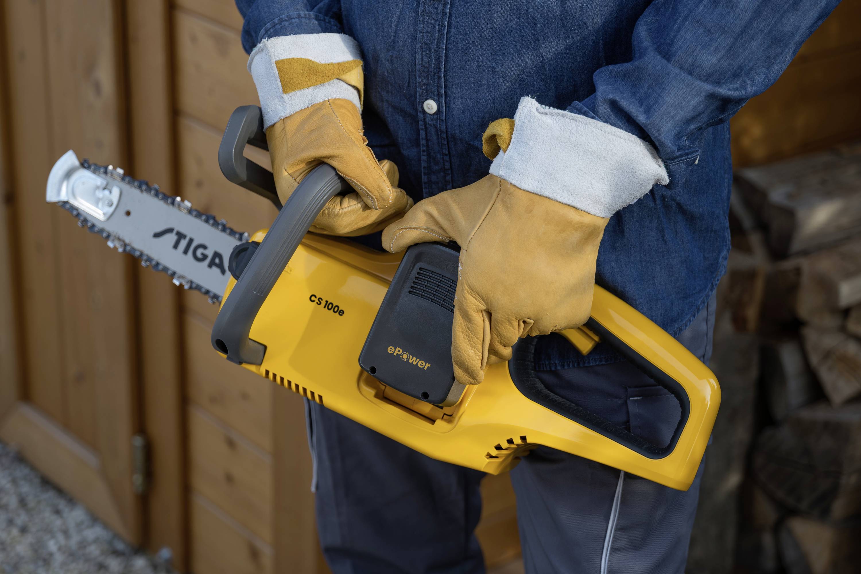 Een persoon houdt een gele kettingzaag met handschoenen, staand voor een houten schuur. De zaag is klaar voor gebruik.