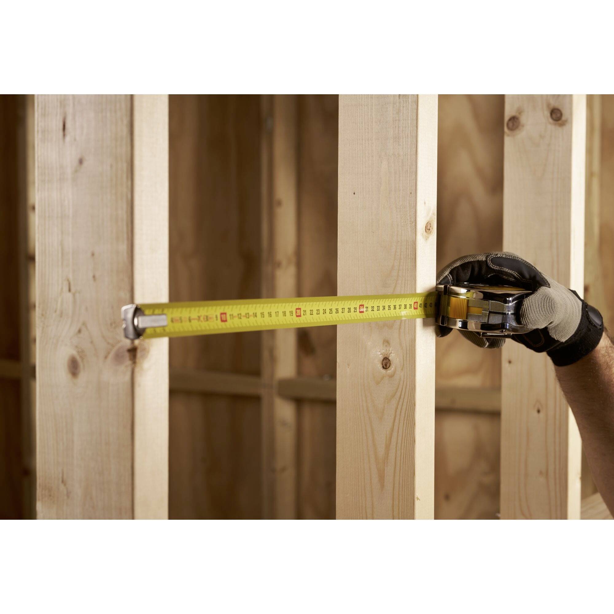 Een gehandschoende hand houdt een meetlint vast dat de afstand tussen twee houten palen meet.