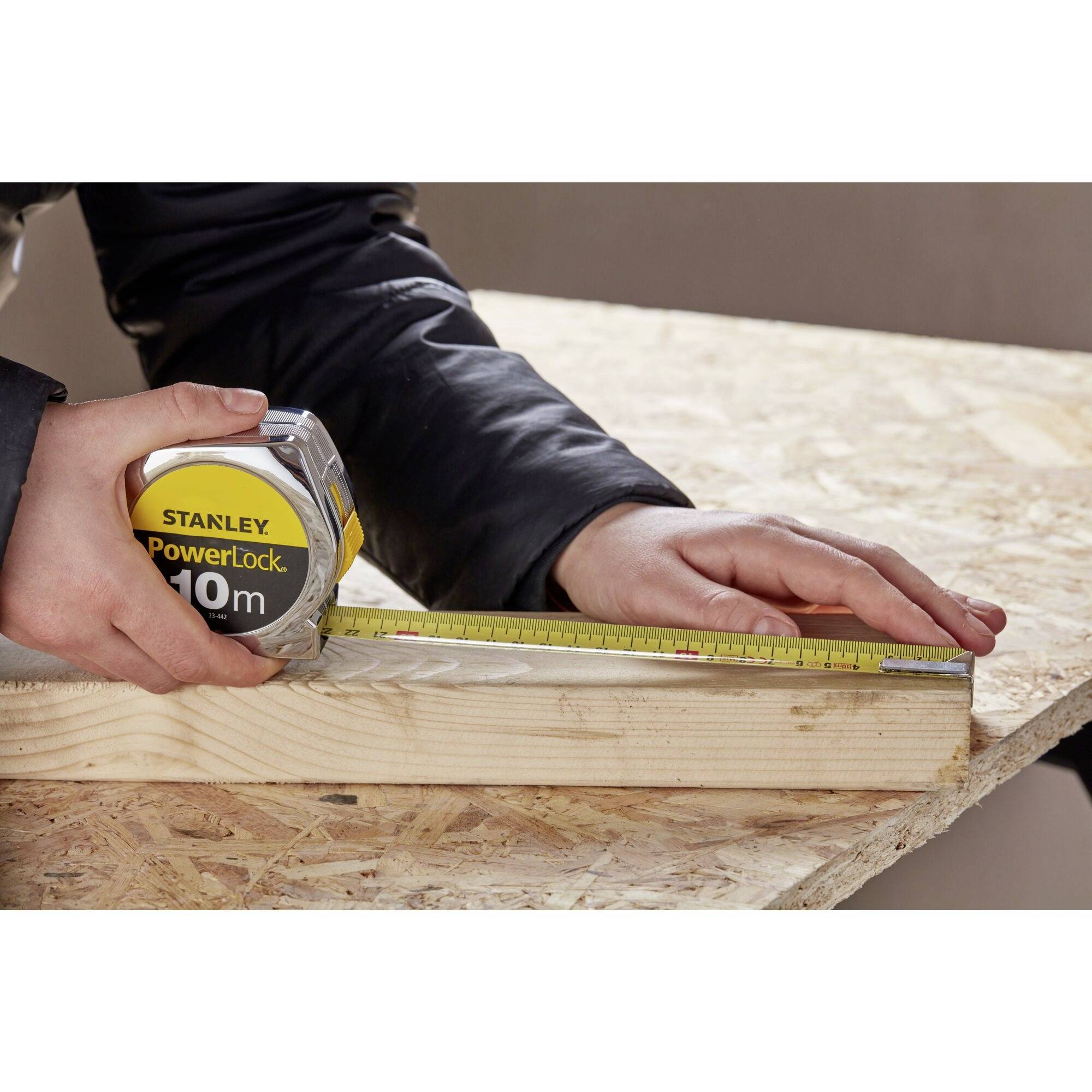 'Twee handen meten een houten plank met een meetlint en lineaal op een multiplex tafel.'