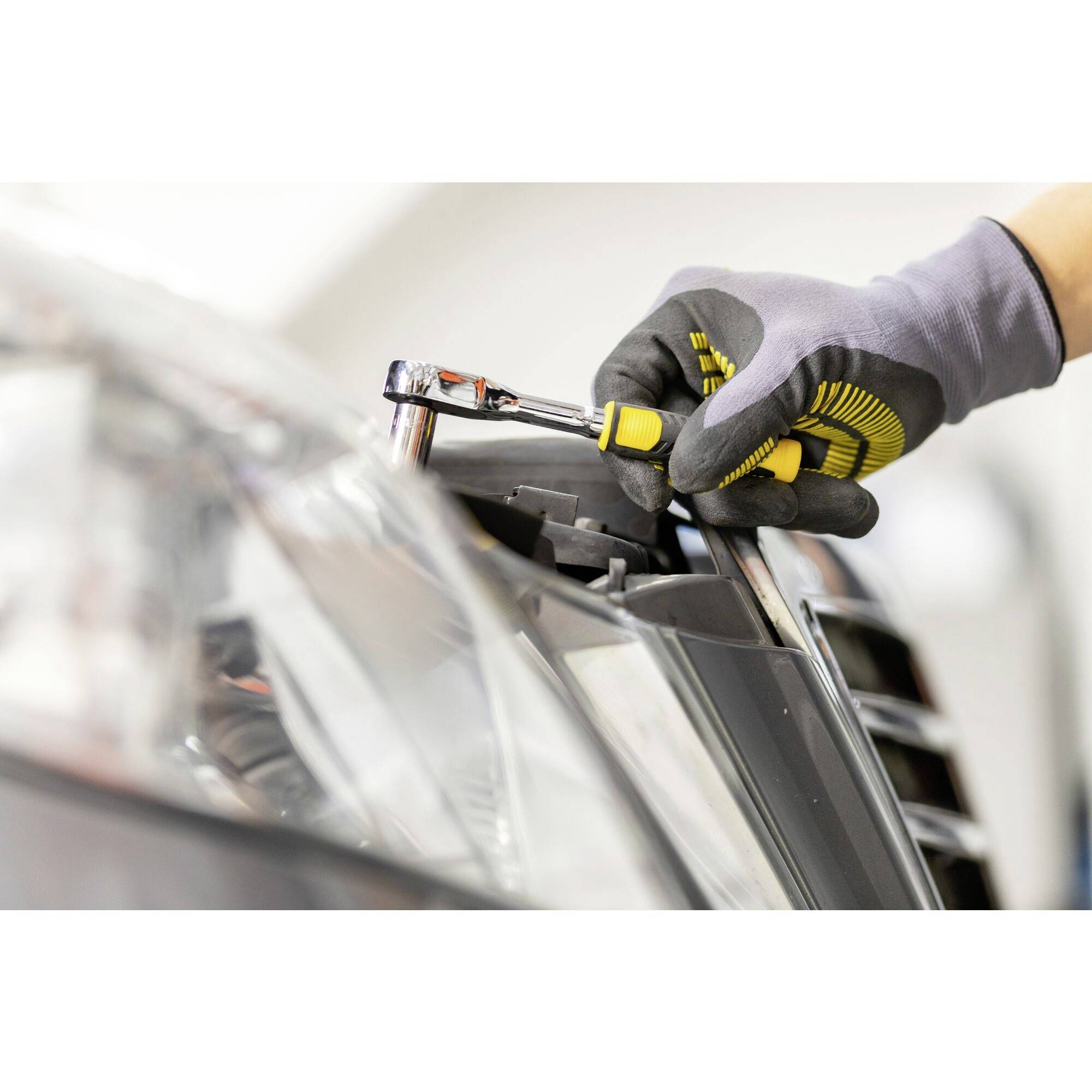 Een persoon met handschoenen draait met een schroevendraaier aan een auto, waarschijnlijk aan een koplamp, in een werkplaats.