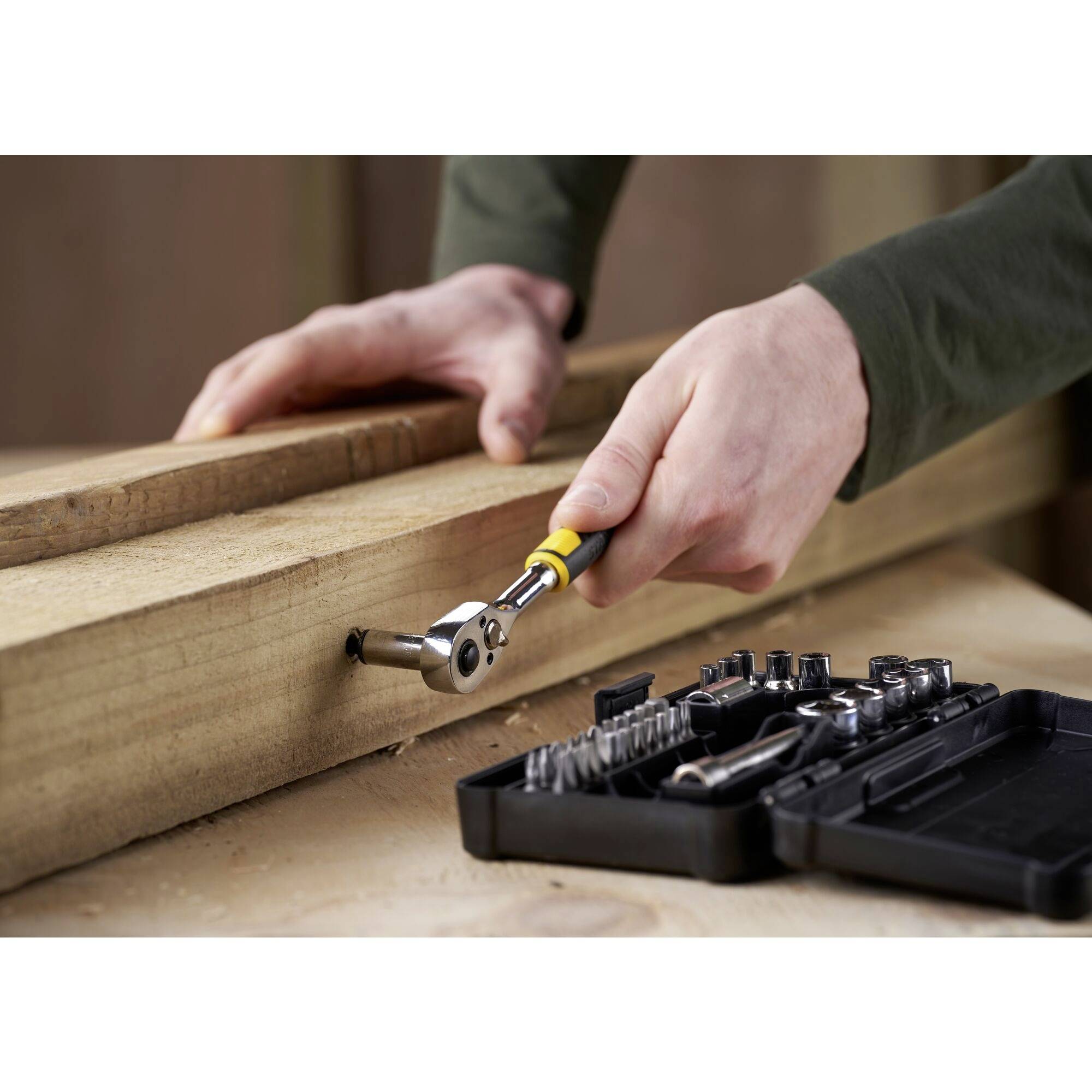 Een persoon gebruikt een ratel om een schroef in een houten plank te draaien. Op de voorgrond ligt een open gereedschapskist met verschillende bitjes.