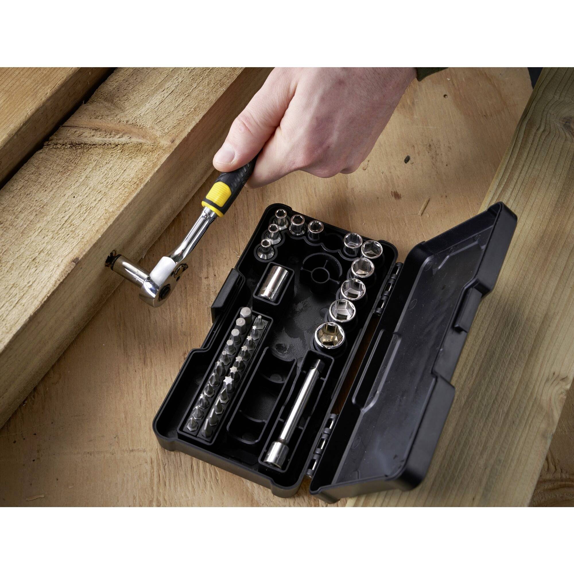 Een hand houdt een ratelgereedschapgreep vast boven een geopende gereedschapskist met verschillende dopsleutels en bits op een houten tafel.